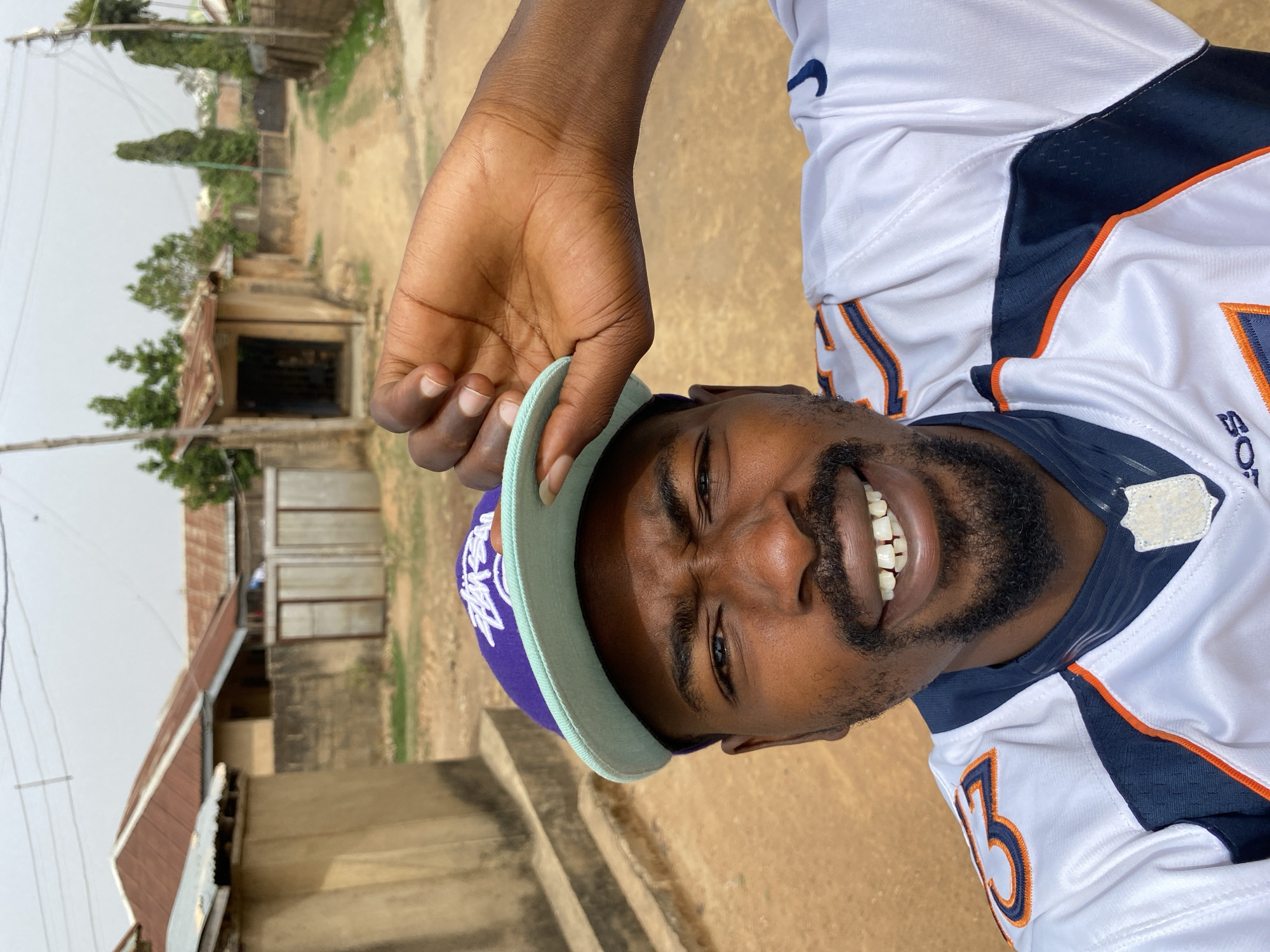 A dark-skinned man stands outside in what appears to be Jos, Nigeria, holding the brim of his purple Los Angeles Lakers baseball cap. He is wearing a white and blue Denver Broncos jersey, number 13. His beard is full and trimmed. He is smiling broadly, showing his teeth. The background shows buildings with corrugated metal roofs and dirt roads, typical of some areas in Jos. The lighting suggests it is daytime.