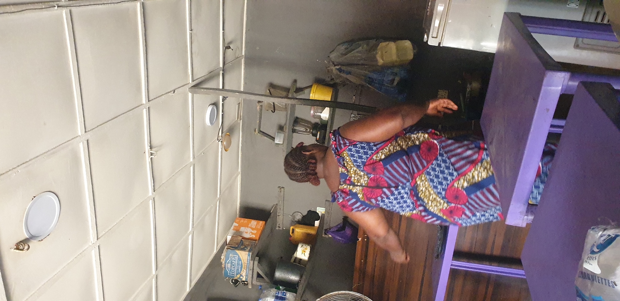 The image shows the back of a woman with braided hair, wearing a colorful patterned dress, in what appears to be a small shop or kitchen. She is standing in front of a wooden counter and a purple table. Behind her, shelves are filled with various items including boxes of "TOPCAFE COFFEE MAX," a yellow jug, a blender, and bags of what seem to be food items. A white refrigerator is visible on the right side of the image. The ceiling is a suspended grid with lights. The environment suggests a commercial setting, possibly a food stall or a small grocery store. The woman appears to be working or attending to something in the area.