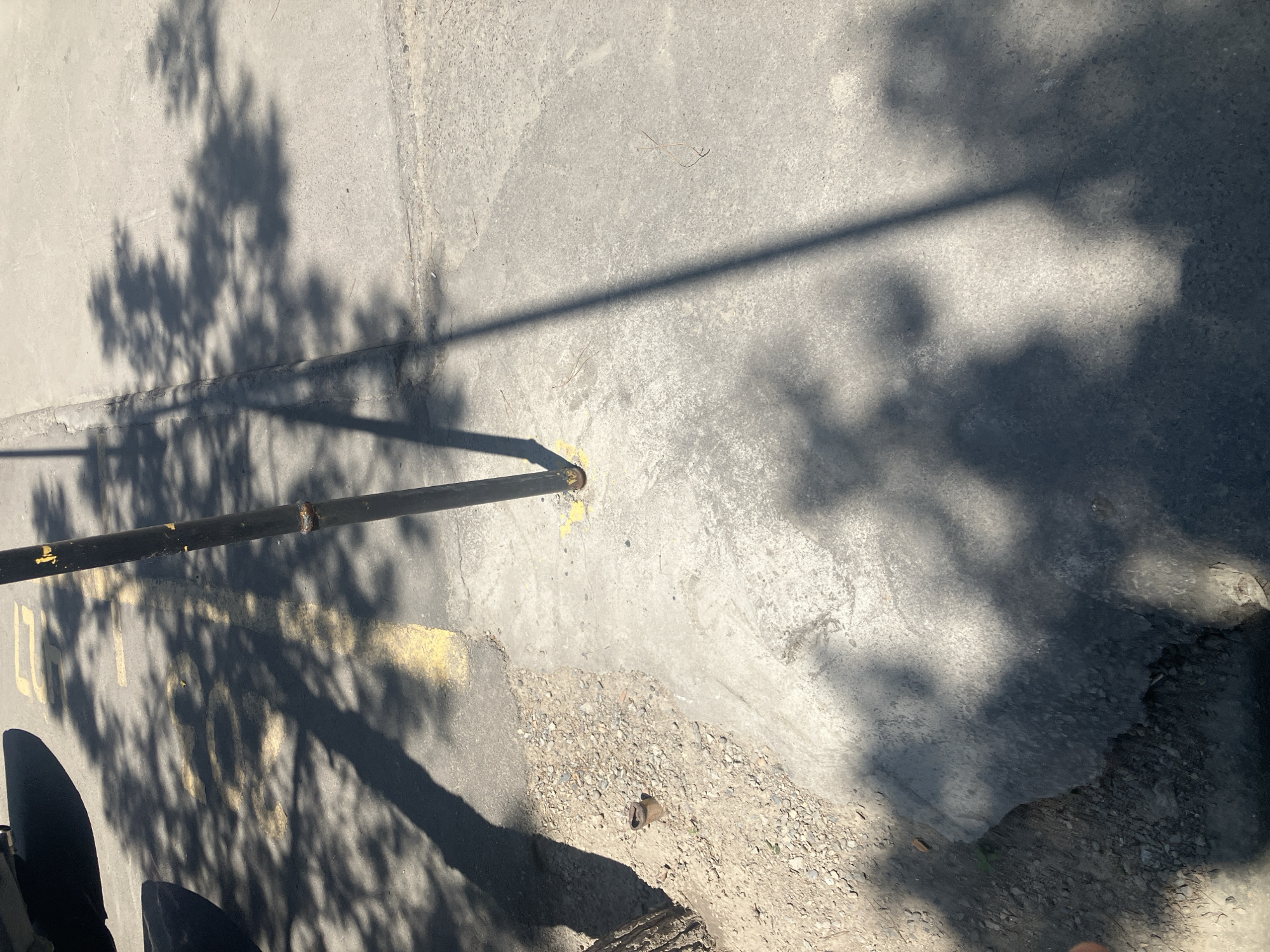 The image depicts a ground-level view of a paved surface, likely a sidewalk or road, casting shadows from unseen foliage above. A black metal pole runs horizontally across the frame, supported by a yellow marker on the ground. Faint yellow markings are also visible on the pavement, with partial numbers such as "1" and "903" discernible. The scene is bathed in bright sunlight, suggesting it is daytime. The ground appears weathered and uneven in places, with small stones and dirt scattered across its surface, particularly near the base of the pole and in a recessed area on the right. The overall impression is of an outdoor urban setting, possibly in Tashkent, Uzbekistan, on a clear day. There are no visible people or animals in the frame, and the focus is on the textures and shadows of the environment.