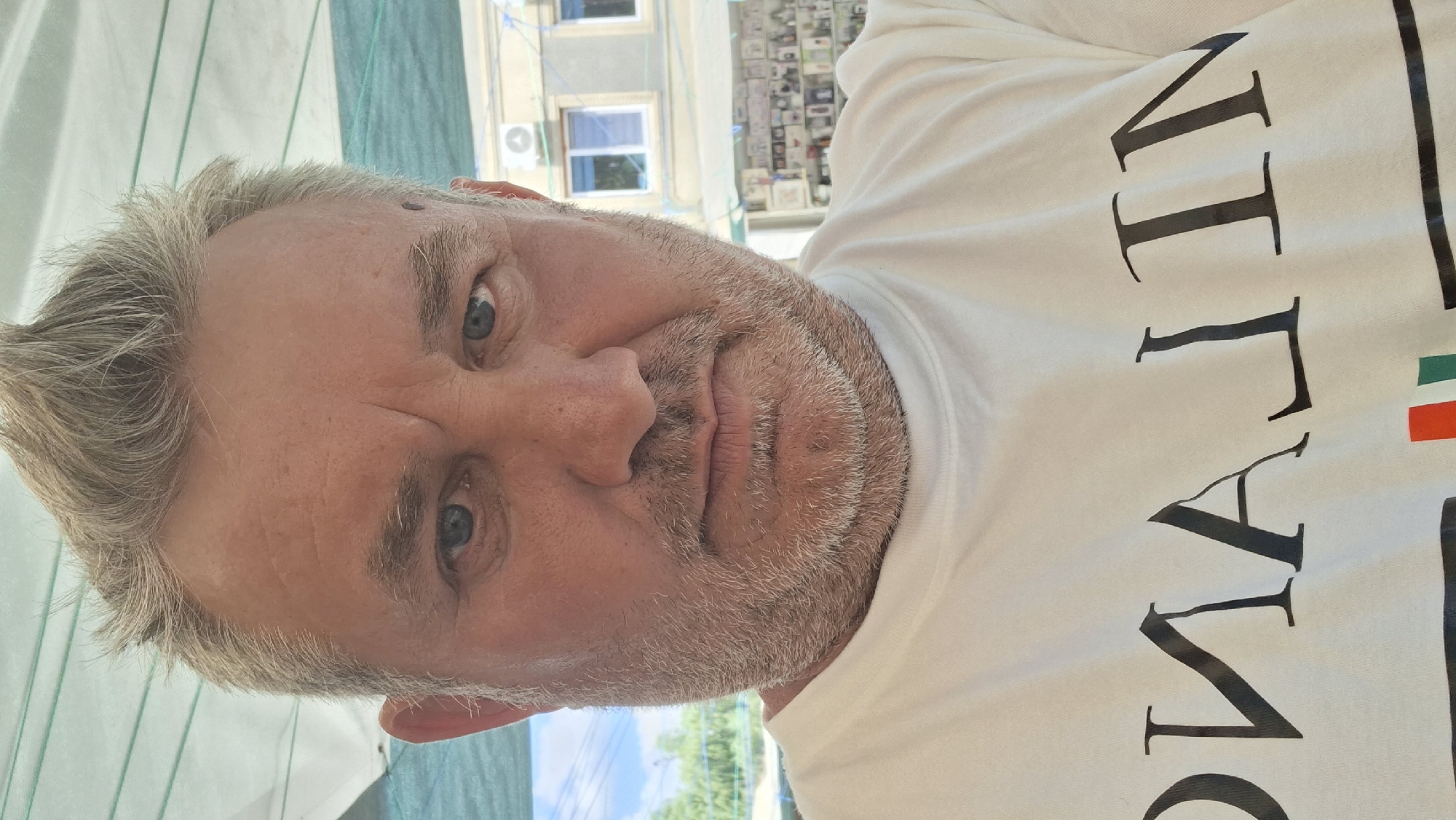 A man with graying, spiky hair and a short, grizzled beard is captured in a close-up, head-and-shoulders selfie. He has blue eyes and is wearing a white t-shirt with large, dark lettering that appears to read "MILANO" and also features the Italian flag's colors. The background suggests an outdoor urban setting with a building visible, possibly shops, and a glimpse of the sky and greenery. The lighting indicates it is daytime.