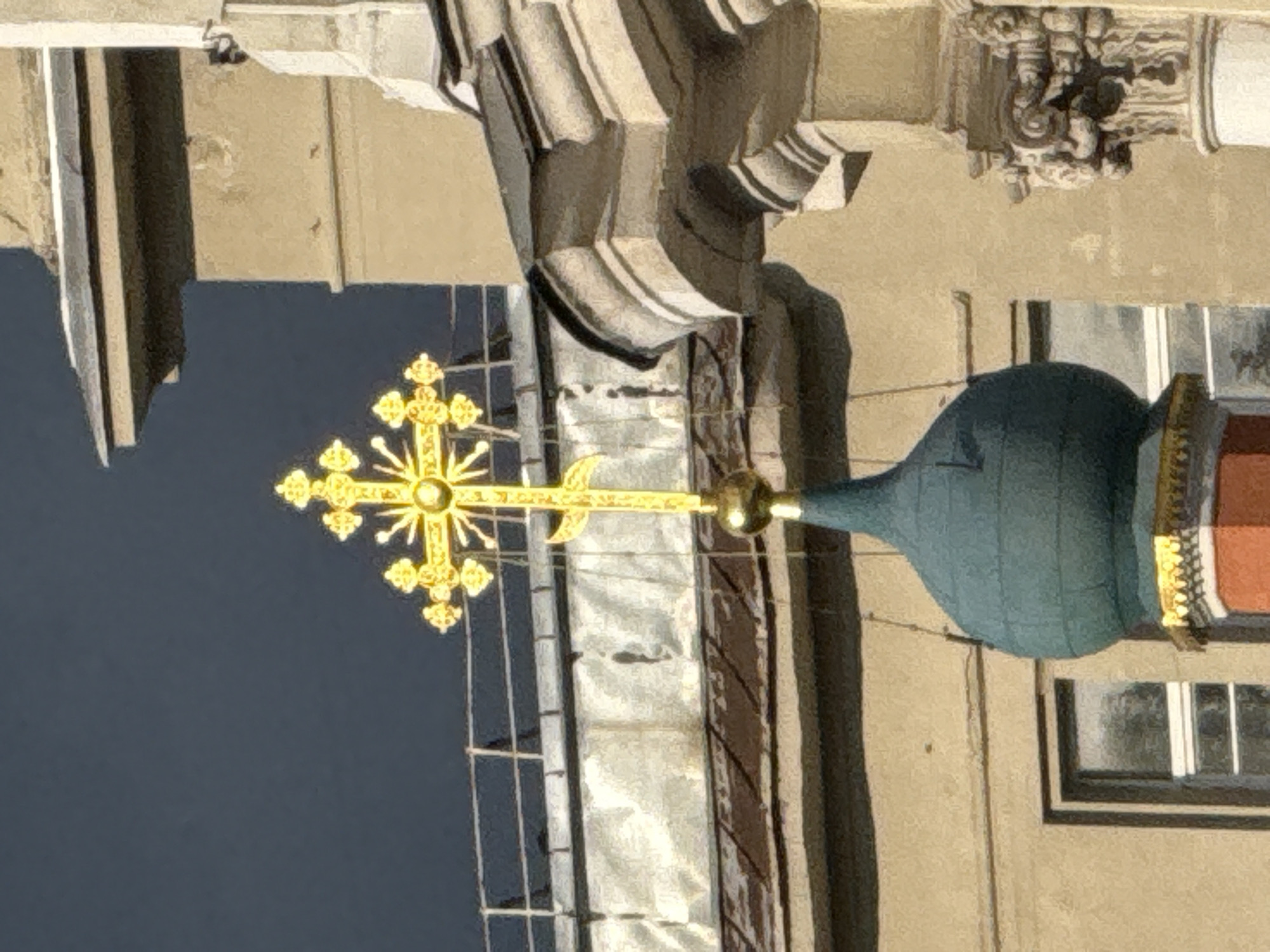 A gold, multi-pointed Orthodox cross stands atop a teal-colored dome, which is itself on top of a red and gold base. The cross is ornate with smaller gold points emanating from the ends of the main cross. The dome is smooth and rounded, and the base features decorative gold trim. Behind the dome and cross, a beige building with visible windows is seen. Part of another building, with elaborate stone carvings, is visible to the right of the cross. The sky in the background is a dark, stormy gray. This appears to be the Saint Nicholas Naval Cathedral in Moscow, Russia.