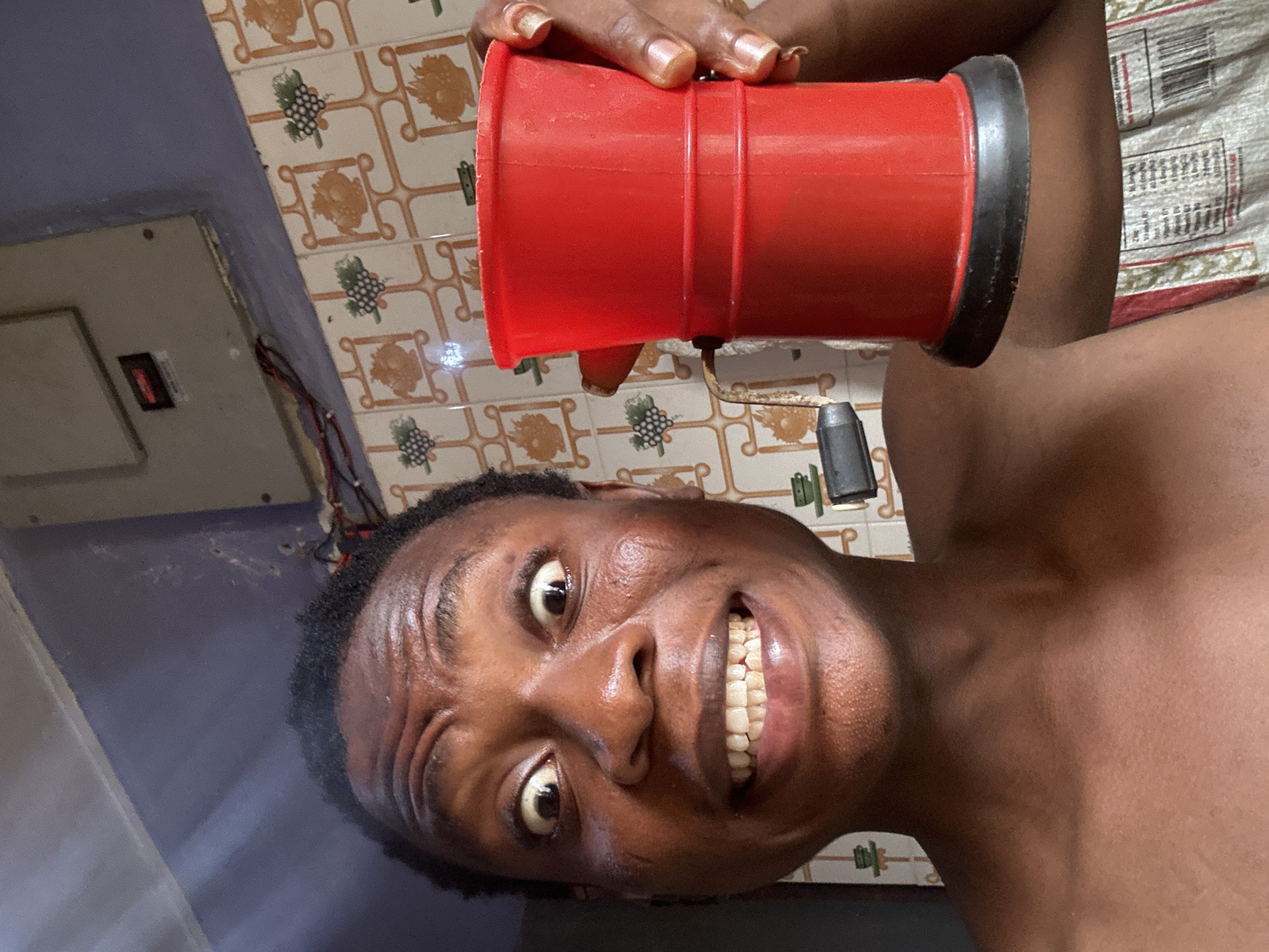 A man is holding a red plastic object with a black rim to his ear, smiling broadly. He appears to be inside a building with tiled walls and a light blue-grey painted area. Electrical wiring and a small electronic panel are visible on the wall behind him. The specific location in the city of Unknown, Unknown is not discernible.