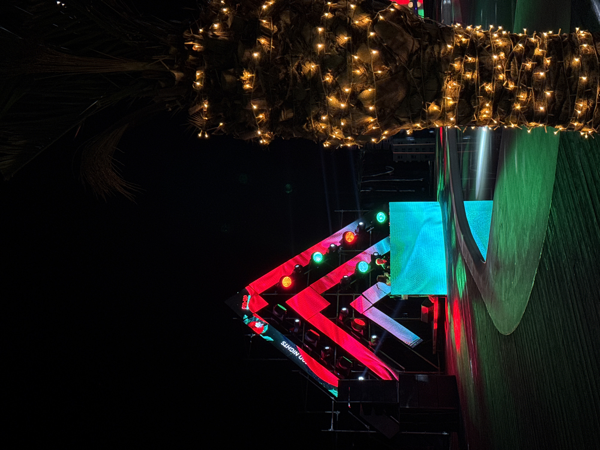 The image depicts a night scene in what is likely Parakar, Armenia. A palm tree trunk, adorned with strings of small, warm-toned lights, occupies the upper portion of the frame. Below, a stage or structure is visible, decorated with brightly lit geometric shapes and LED screens displaying various colors. The structure's design includes red, green, and blue lights, forming a chevron pattern. A portion of the structure shows text "GOOD NIGHTS". The rest of the scene fades into darkness.