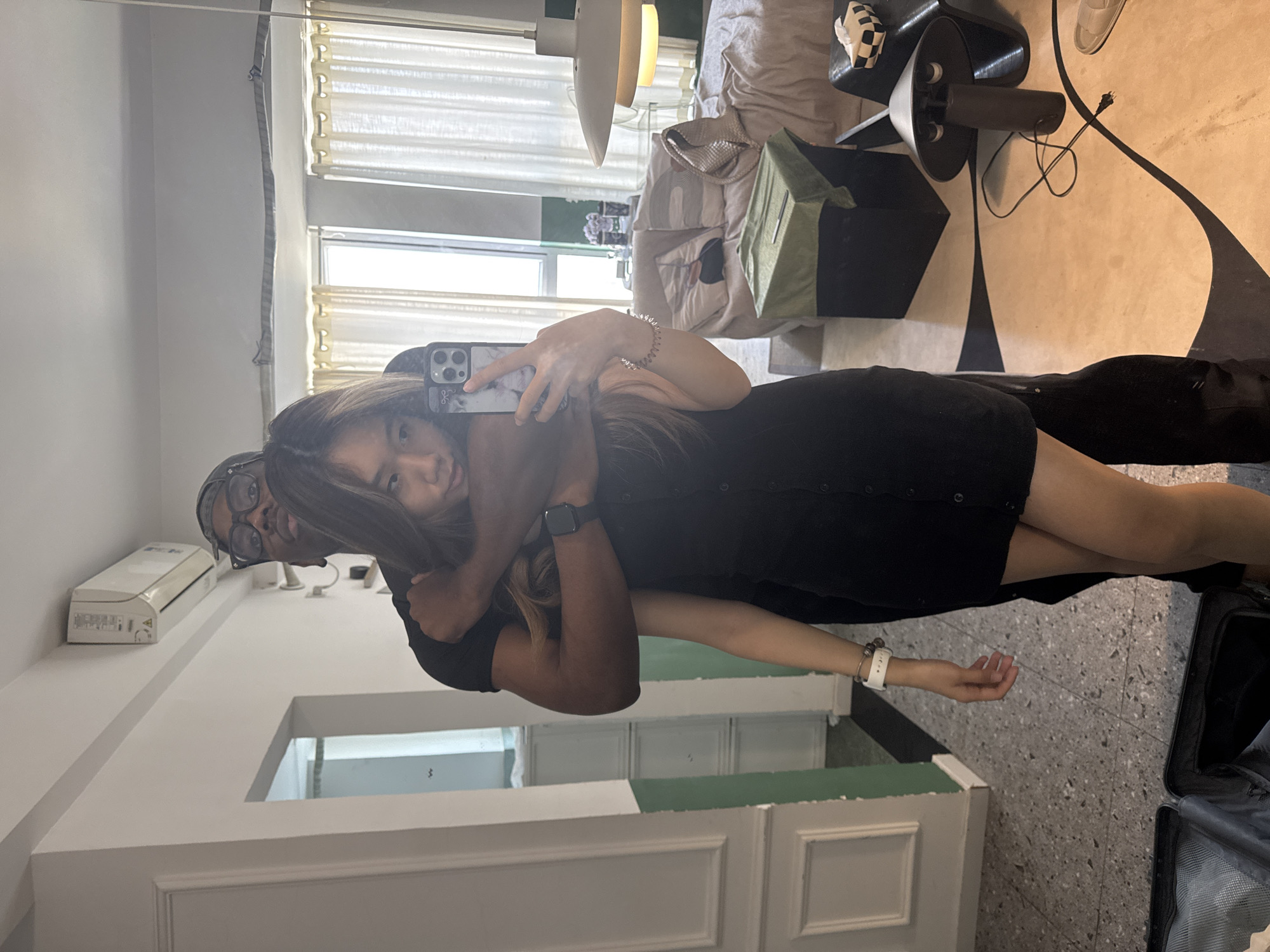 A woman in a black dress takes a selfie with a man embracing her. The location appears to be inside a residential building. A luggage bag is on the right, implying a trip. The presence of an air conditioner unit suggests a modern interior in Liangjiang, China.