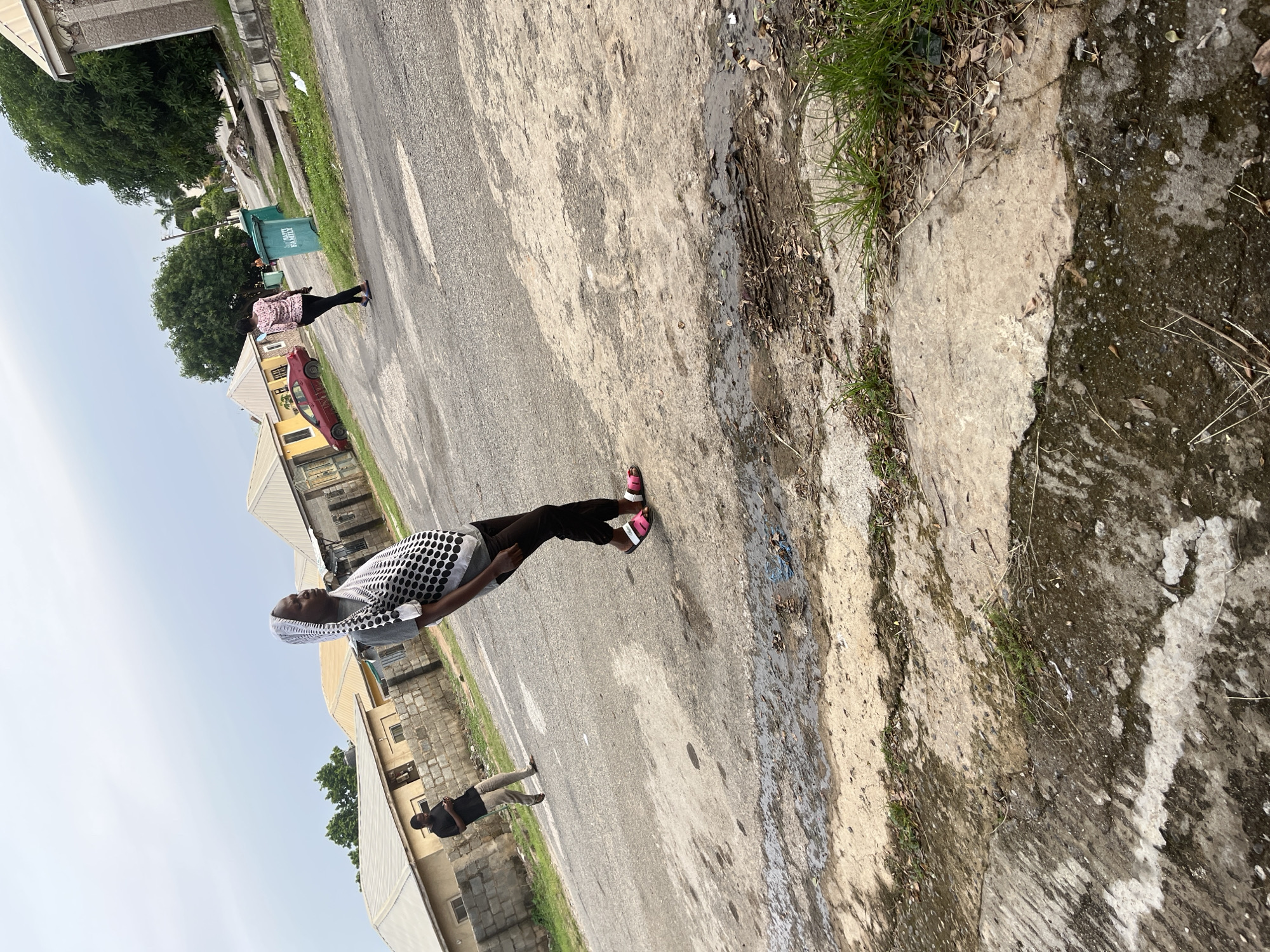 A medium-shot, outdoor, eye-level, full-color photograph captures a residential street scene in Abuja, Nigeria. The sky is overcast, with diffused light illuminating the scene. In the foreground, a person wearing a white headscarf with a black and white dotted pattern over a grey t-shirt and black pants stands with their left hip out, looking to their left. They are wearing pink and white slide sandals. To the left and further back, a person in a black t-shirt and light brown pants is walking while looking at their phone. In the middle ground, a woman in a pink patterned shirt and black pants is walking away from the camera. Behind her, a green trash bin with "HAPPY FAMILY" written on it is visible, as is a red car parked on the side of the street. The street is paved with asphalt and shows signs of wear, with patches of dirt and debris. On the left side of the street, low-rise residential buildings with grey concrete block walls and silver-colored roofs are present. Trees with green foliage are scattered throughout the scene. The overall impression is of a quiet, everyday street in a residential area.