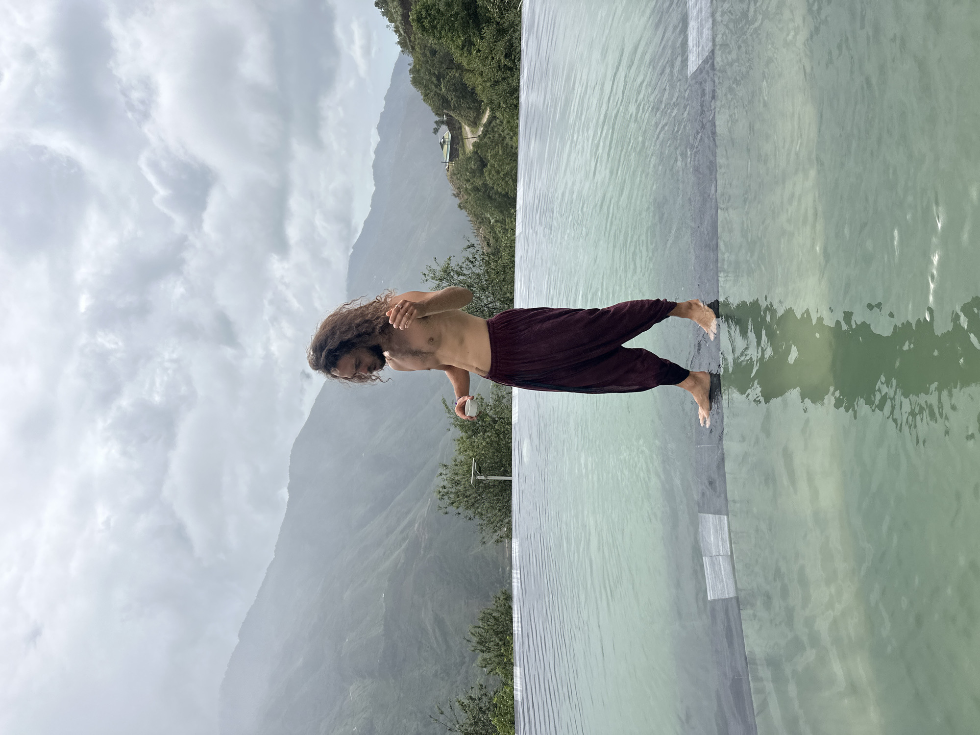 A man with long, curly brown hair and a beard stands barefoot at the edge of an infinity pool, holding a small white cup in his right hand. He is shirtless and wearing loose, maroon pants. The pool's water is a pale green, and it reflects the overcast sky and the surrounding mountainous landscape. In the background, lush green mountains rise under a cloudy sky, with hints of a road winding up one of the slopes and a small building visible on the hillside. The location is identified as Ta Xua, Vietnam.