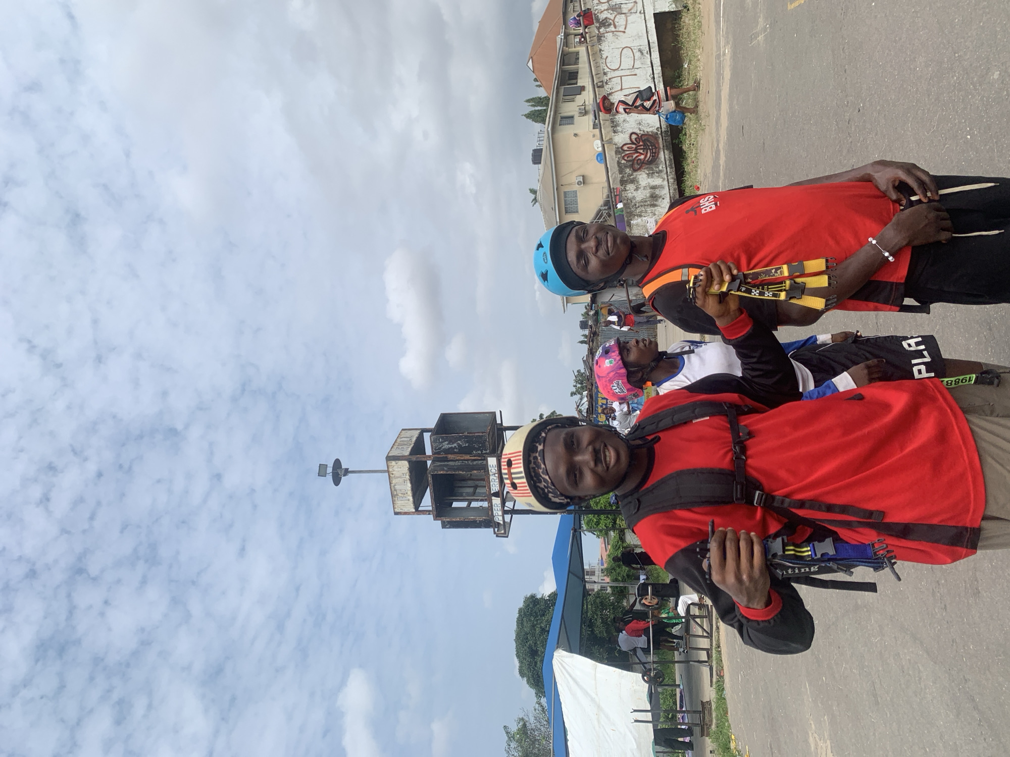 Two young Black men are standing outdoors in Alagbede, Nigeria, with a cloudy sky overhead. The man on the left is wearing a red long-sleeved shirt with black sleeves, a black backpack, and a tan and black patterned helmet. He is holding a set of black straps with yellow and blue accents. The man on the right is wearing a red sleeveless vest over a black shirt, black pants, and a blue helmet. He is also wearing a black backpack. They appear to be prepared for an activity. In the background, there is a large, weathered, multi-level structure with what appears to be speakers or ventilation units. Further in the background, there are buildings, some with graffiti, and other people, suggesting a public or communal space. A white tent or canopy is visible to the left, and some exercise equipment, including a weight bench, is present in the middle ground.