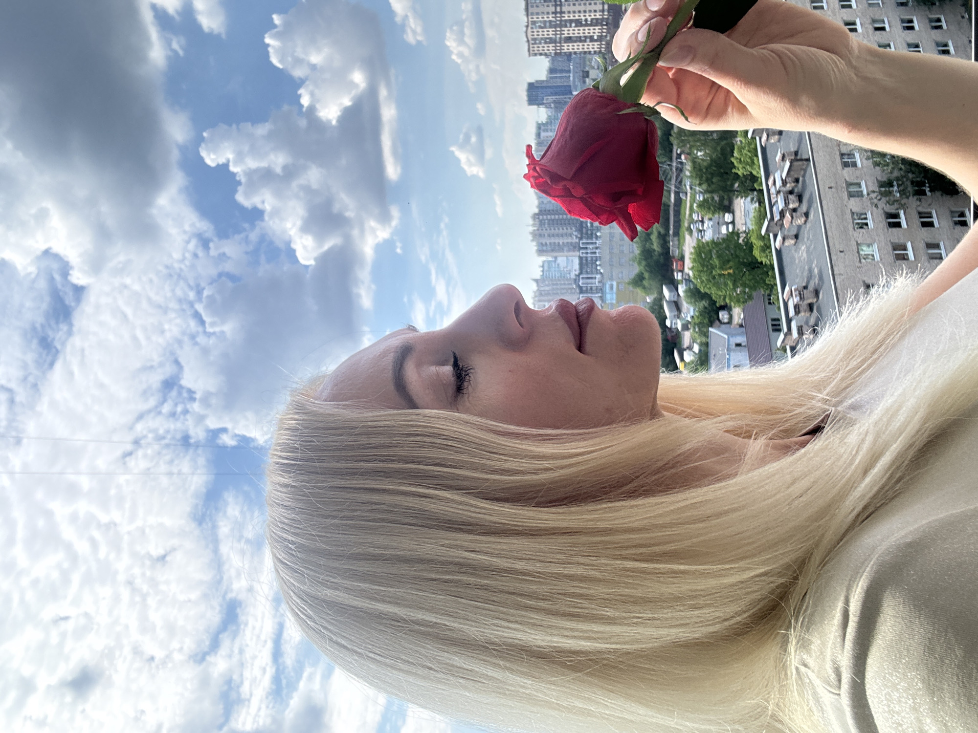 The image displays a woman in profile, positioned on the right side of the frame, gazing upwards towards a vibrant red rose. She has long, straight, very light blonde hair, and her eyes are gently closed, suggesting a moment of tranquility or enjoyment as she brings the rose closer to her nose, as if to inhale its fragrance. Her lips are slightly parted, and her facial features appear relaxed. She is wearing a light-colored garment, partially visible.
The background features a bright blue sky filled with numerous white and grey fluffy clouds, indicating a clear, sunny day. Below the horizon line, an urban landscape extends, characterized by tall, multi-story residential buildings, some appearing as grey concrete structures with multiple windows. Green trees are interspersed among the buildings, adding a touch of nature to the city view. The perspective suggests the photo was taken from a significant height, possibly a balcony or a high window overlooking the cityscape. No visible text is present in the image. The overall mood is peaceful and contemplative. The architectural style of the buildings is consistent with urban residential areas in Russia, aligning with the provided context.