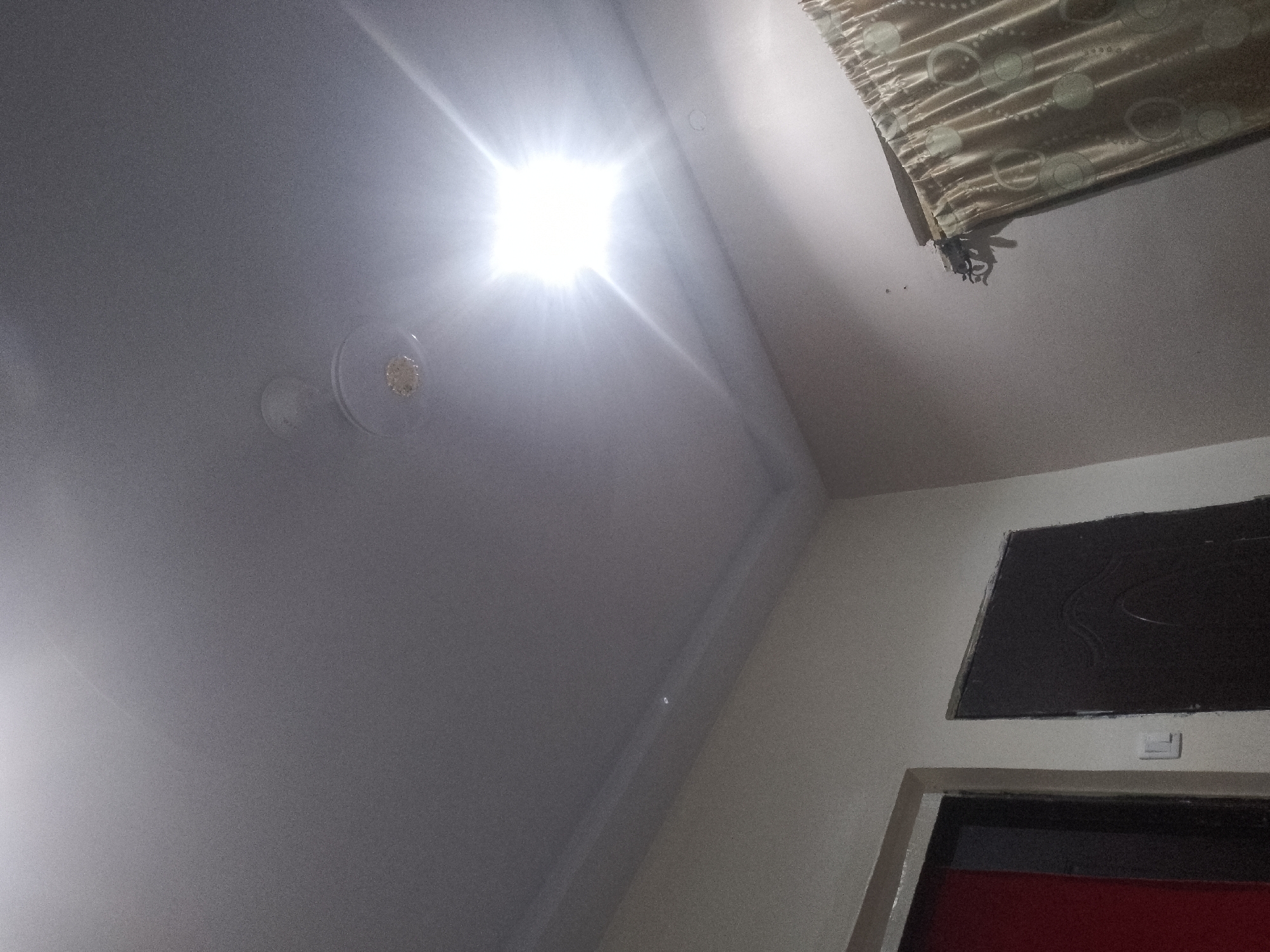 An indoor, low-angle shot of a ceiling shows a bright overhead light fixture reflecting off the white surface, with a ceiling fan visible below it. A tan and beige patterned curtain is draped in the upper right corner. Below the curtain, a small, dark object, possibly a spider, hangs precariously. Further down the wall, a dark rectangular ventilation grate is mounted above an open doorway, revealing a dark interior with a red element visible at the bottom. The lighting in the scene is harsh and uneven, with significant glare from the overhead light.