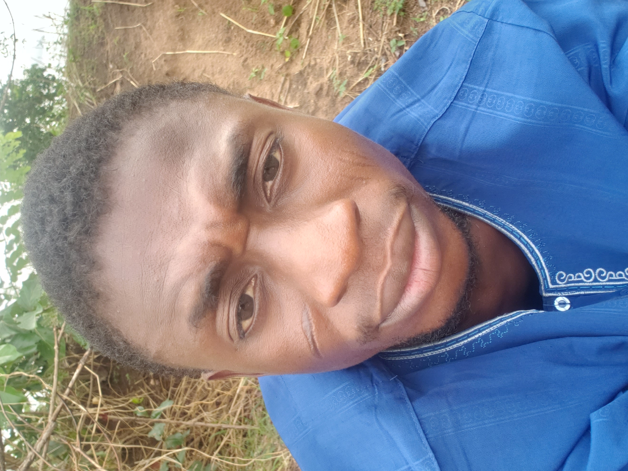 A close-up, eye-level view of a dark-skinned man wearing a blue shirt with patterned collar and sleeves, taken outdoors in daylight. The man has short, dark hair and a beard. He is looking slightly down at the camera. There appears to be a small scar on his left cheek. The background shows dry earth and some sparse vegetation. The specific location in Adankolo, Nigeria cannot be confirmed with only this image.