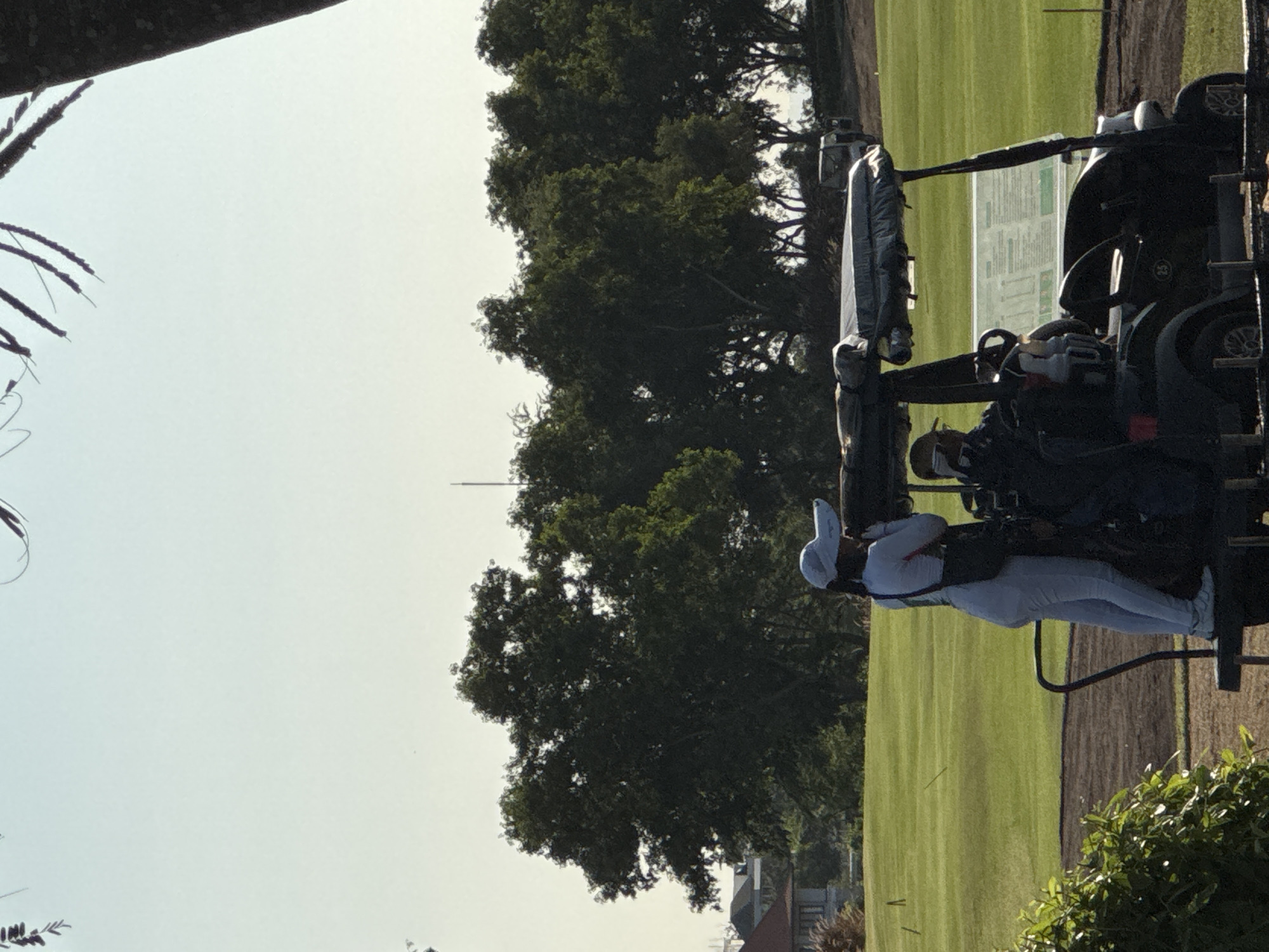 A person in a white outfit stands next to a golf cart on a green lawn in Kuta Selatan, Indonesia. Trees line the background of the image, and the sky is bright. The person is seen interacting with the golf cart.