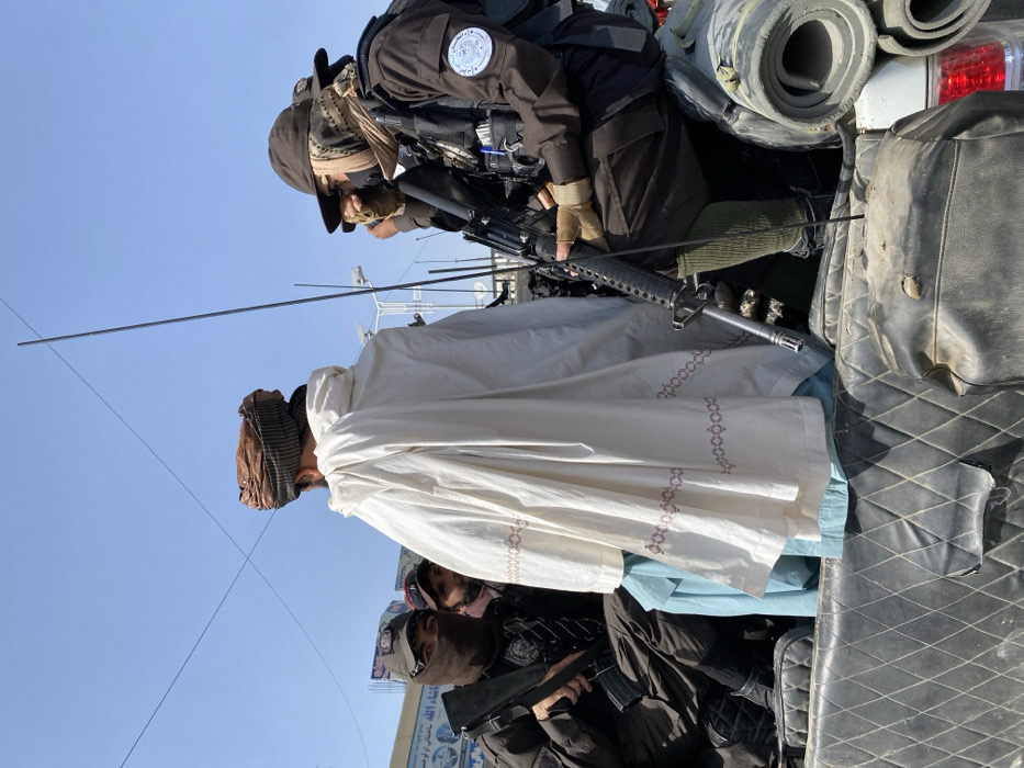 The image captures a daylight scene on an urban street in Kandahar, Afghanistan. Three prominent male figures are situated within the bed of a utility vehicle, likely a pickup truck, which features a black, diamond-quilted vinyl lining on its inner walls.
The central figure, viewed from the back, wears a light brown turban, a white outer garment with brown patterned trim along the bottom, and a light blue tunic beneath. To his immediate left, a male in a dark uniform wears a black balaclava obscuring most of his face, a dark cap, and dark tactical gear with a dark sleeve patch. He holds a dark assault rifle (M4/AR-15 variant) across his lap, facing towards the viewer.
To the central figure's right, another male in a dark uniform wears a brown wide-brimmed hat, a tactical scarf partially covering his face, and dark tactical gear. His right sleeve bears a circular patch with Farsi script, which appears to read "Islamic Emirate of Afghanistan." He holds a dark assault rifle (M4/AR-15 variant) with his right hand resting on the handguard, while his left hand is raised towards his face. He wears light-colored gloves and olive-green leg coverings.
Two thin, dark vertical antennae extend upwards from the vehicle bed. Rolled-up mats or bedding are visible on the right side of the vehicle bed. In the background, under a clear blue sky, buildings with signage and overhead power lines indicate an urban environment. A partially visible male with a dark cap is present behind the central figure and to the left.