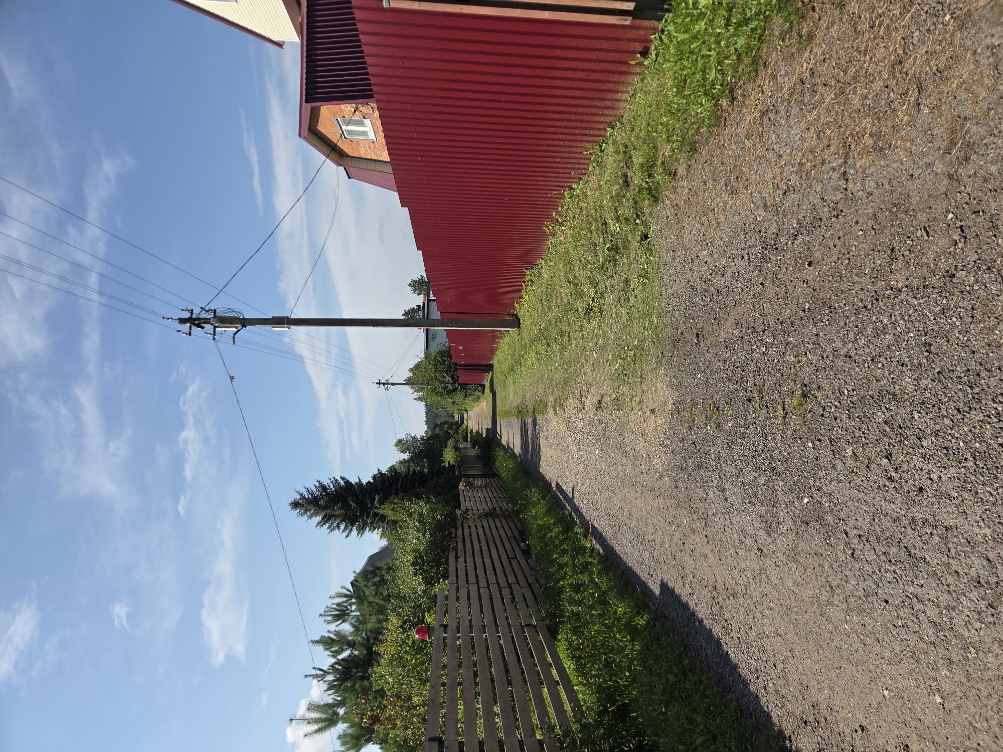 The image depicts a rural dirt road in Frolovo, Russia, flanked by fences and vegetation under a clear blue sky with scattered clouds. On the left side of the road, a light-colored wooden plank fence stretches into the distance, with lush green grass growing along its base. Beyond this fence, trees and bushes form a dense natural barrier. A red flashing light is visible atop a pole behind the wooden fence. On the right side of the road, a tall, corrugated red metal fence dominates the view, with a light-colored house partially visible behind it. A telephone pole with multiple wires is situated near the red fence, casting a shadow on the road. The road itself is unpaved and composed of gravel and dirt, showing tire tracks and some patches of dry grass. The overall scene suggests a quiet, rural setting on a sunny day.