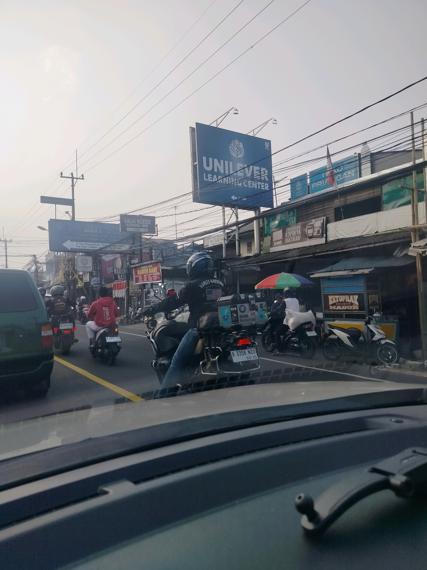 The image shows a traffic scene in Megamendung, Indonesia. A variety of vehicles, including cars and motorcycles, are visible on a road lined with shops and advertisements. A large blue billboard for "UNILEVER LEARNING CENTER" is prominent in the background. Several people are riding motorcycles, some wearing helmets and casual clothing. One person is wearing a red hoodie and white pants. The environment appears to be a busy urban or semi-urban area with overhead electrical wires and street signs. The overall lighting suggests it is daytime.