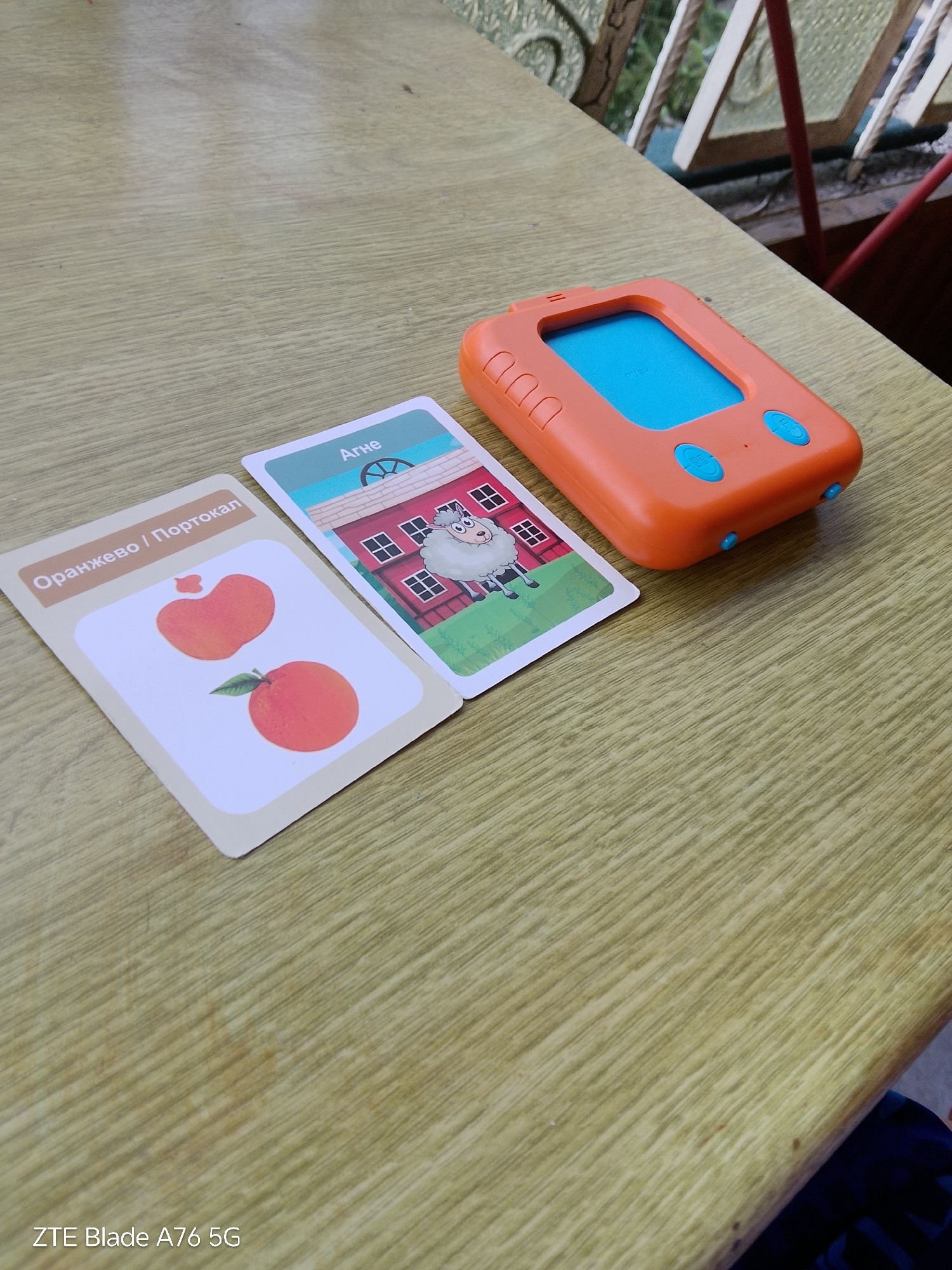 An orange electronic toy, shaped like a small handheld device with a blue screen and two blue buttons, sits on a light-colored wooden table. Two flashcards are placed next to the toy. The first card, on the left, features two images of oranges, one with a leaf attached. The text on this card reads "Оранжевo / Портокал" (Orange / Mandarin). The second card, to the right of the first, depicts a cartoonish red barn with windows and a sheep standing in front of it. The text on this card reads "Агне" (Lamb). The scene is indoors, likely on a table, with a hint of a balcony railing and greenery visible in the background, suggesting an outdoor view from a window. The lighting appears to be natural, indicating daytime. No people are visible in the frame. The bottom left corner of the image displays the text "ZTE Blade A76 5G".