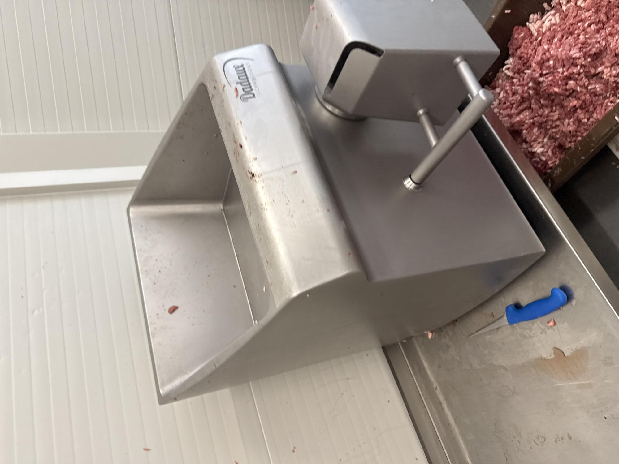 The image shows a stainless steel meat grinder, possibly a Dadaux brand, situated on a stainless steel counter with a blue-handled knife nearby. The area appears to be a commercial food processing or butcher shop, judging by the presence of ground meat in a container adjacent to the grinder. The background consists of a white, panelled wall. No people are visible. There is no information in the image to suggest the location is Stara Zagora, Bulgaria.