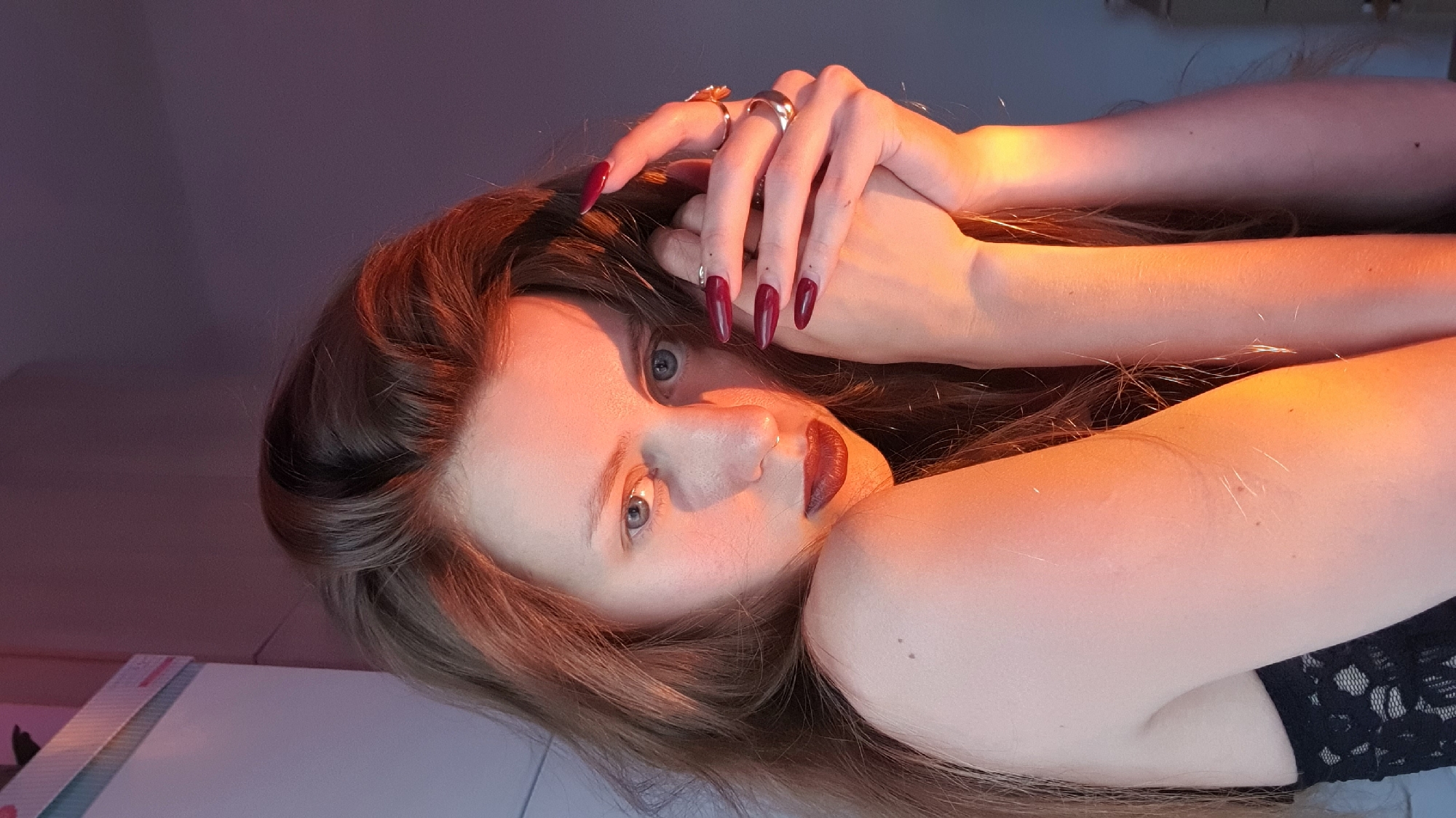 A young, light-skinned woman is lying on her side in a dimly lit interior. She has long, light brown hair and blue eyes. Her lips are painted with dark red lipstick and her fingernails are long and painted dark red. She is wearing a black lace top. Her hands are raised, with fingers interlaced above her head. She wears several rings on her fingers. A strong orange/red light illuminates her face and arms, while a purple light is visible in the background. The location and city of Bar, Montenegro is not discernible from the image.