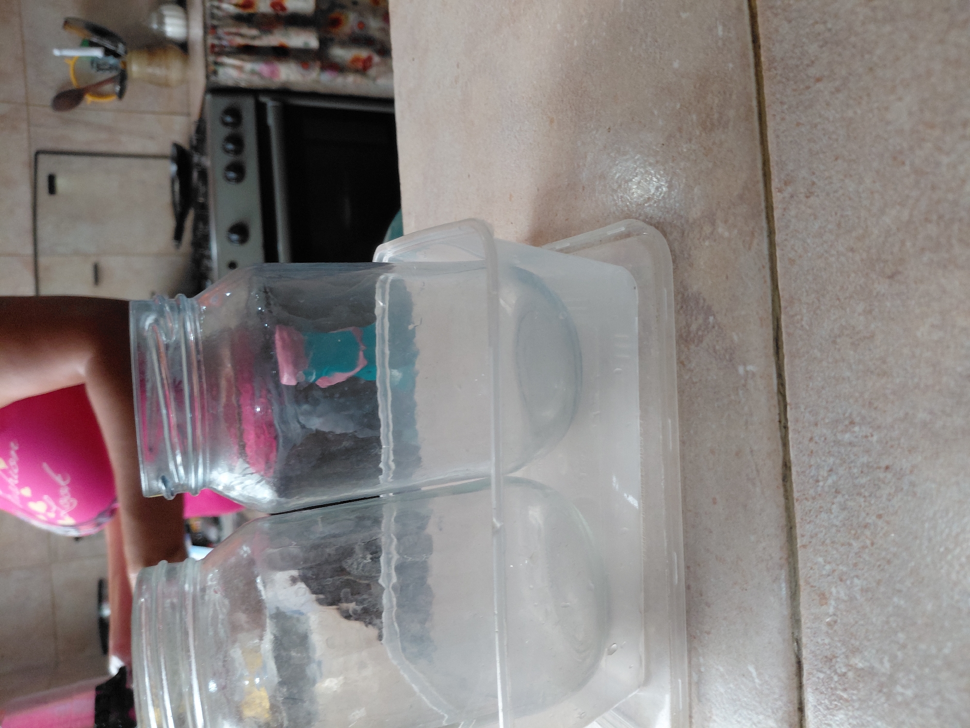 This image depicts an indoor scene, likely a kitchen in Santa Ana, Venezuela, with a focus on storage containers. The view is oriented vertically, showing elements that would typically be horizontal (floor/counter) as vertical.
In the foreground, two clear glass jars with threaded necks are visible, placed upright within a larger, transparent plastic rectangular organizer or tray. Faint colorful material, appearing blue and pink, can be seen inside the upper glass jar.
To the left, a person is partially visible, primarily their right arm and a portion of their torso, wearing bright pink attire. Faint white text, including words like "Love," is discernible on the pink fabric. The lower part of their legs and dark footwear are also seen at the bottom left.
The background reveals more of the kitchen environment. A silver-colored stove or oven with dark control knobs is situated on the left side of the frame. Above it, various kitchen utensils, such as wooden spoons and spatulas, are neatly stored in a holder. The floor and adjacent wall are covered with light beige or cream-colored tiles, separated by dark grout lines. The scene suggests a moment of domestic activity, possibly involving organization or preparation in the kitchen.