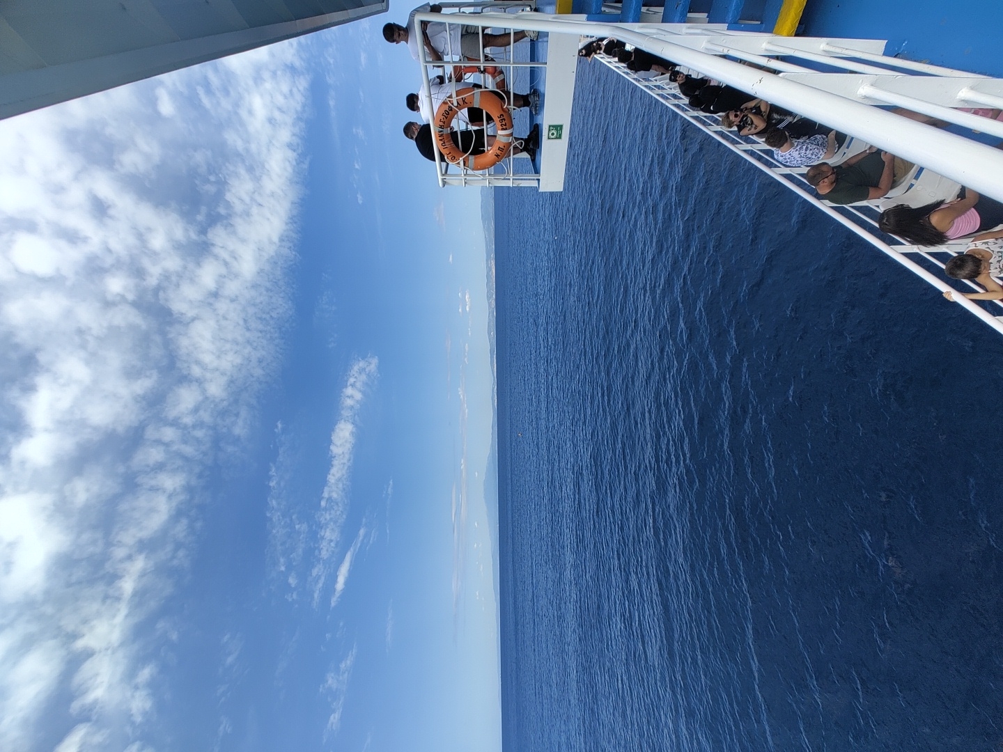 An exterior, high-angle, eye-level shot captures a ferry sailing on the deep blue Aegean Sea. The ferry’s deck railing, painted white and blue, spans the right side of the frame. On the upper deck, four individuals stand near a life preserver, their backs to the camera. A life preserver, orange with a white stripe and the number "255" printed on it, is secured to the railing. On a lower deck, visible in the lower right quadrant, several passengers are seated on benches, facing away from the sea. The horizon displays a distant, faint landmass under a bright blue sky adorned with scattered white clouds. The sea's surface is calm with gentle ripples. The scene is likely in the waters surrounding Thassos, Greece, a common ferry route.