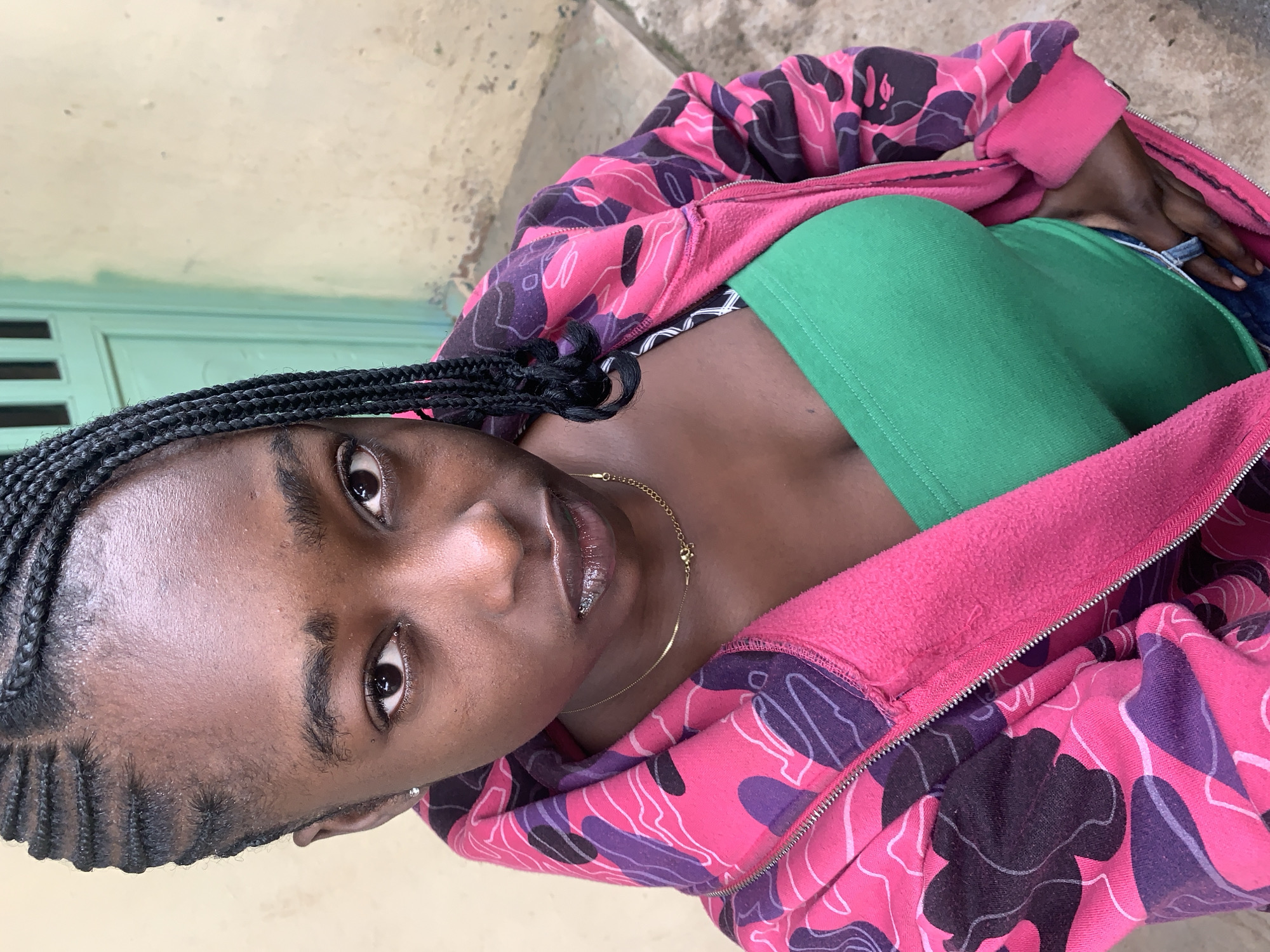 A young Black woman, with braided hair, is pictured outdoors. She is wearing a pink and purple camouflage print hooded jacket over a green tube top and blue jeans. Her gaze is directed towards the camera, and her lips are slightly parted, revealing what appears to be braces. A thin gold chain is visible around her neck. The background consists of a textured, light-colored wall and a teal-colored door. The scene appears to be in a residential or urban setting in Rafin Par, Nigeria.