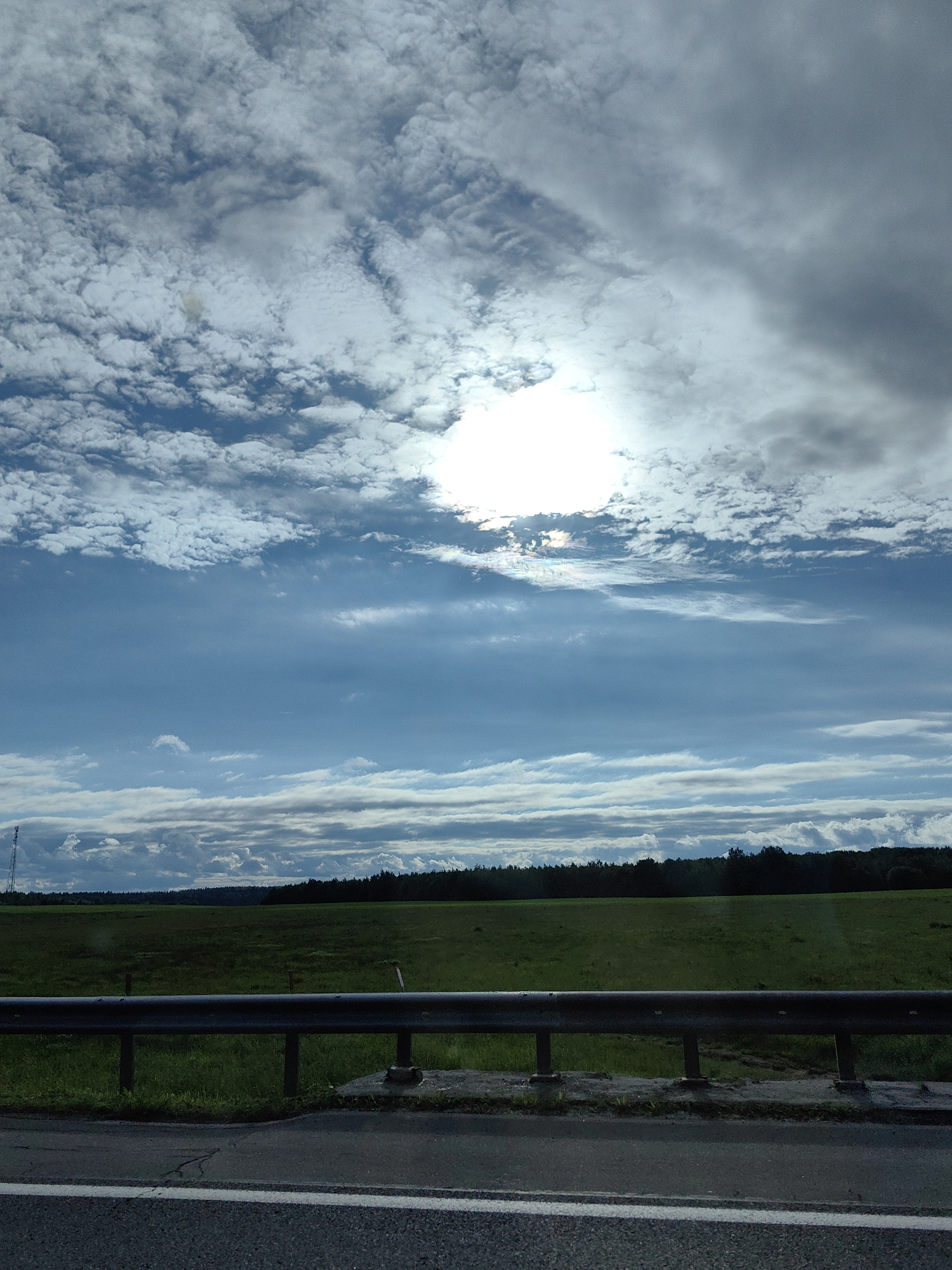 The image depicts a rural landscape in Негарэльскі сельскі Савет, Belarus, observed from the perspective of a vehicle. The sky occupies the majority of the frame, presenting a complex cloudscape. In the upper left, dense, fragmented white cumulus clouds are visible against a lighter blue sky. Towards the upper right, the clouds transition into a darker, more uniform grey overcast, suggesting denser cloud cover. The sun is prominently positioned near the top-center, partially obscured by bright white clouds, creating a strong glare and visible light diffraction/iridescence, with subtle rainbow hues around its edges. A band of clearer, deep blue sky stretches horizontally across the middle, interspersed with thin, elongated white cloud formations. Lower in the sky, nearer the horizon, multiple layers of grey and white clouds are present.
The midground features a vast expanse of dark green fields, which appear somewhat muted due to the mixed lighting conditions. A dense, dark green forest line forms the horizon, separating the fields from the sky. On the far left, a tall, slender communication tower or similar structure is partially visible against the sky, rising above the distant trees.
The foreground is dominated by a dark grey metal guardrail, extending horizontally across the lower portion of the image, indicating the edge of a road or highway. Below the guardrail, a narrow strip of green grass and some darker ground material are visible. The very bottom of the frame shows a section of asphalt road, marked with a clear, solid white line on its left edge. No people, animals, or specific actions are discernible within the frame. The scene conveys a blend of bright sunlight and overcast conditions over an open landscape.