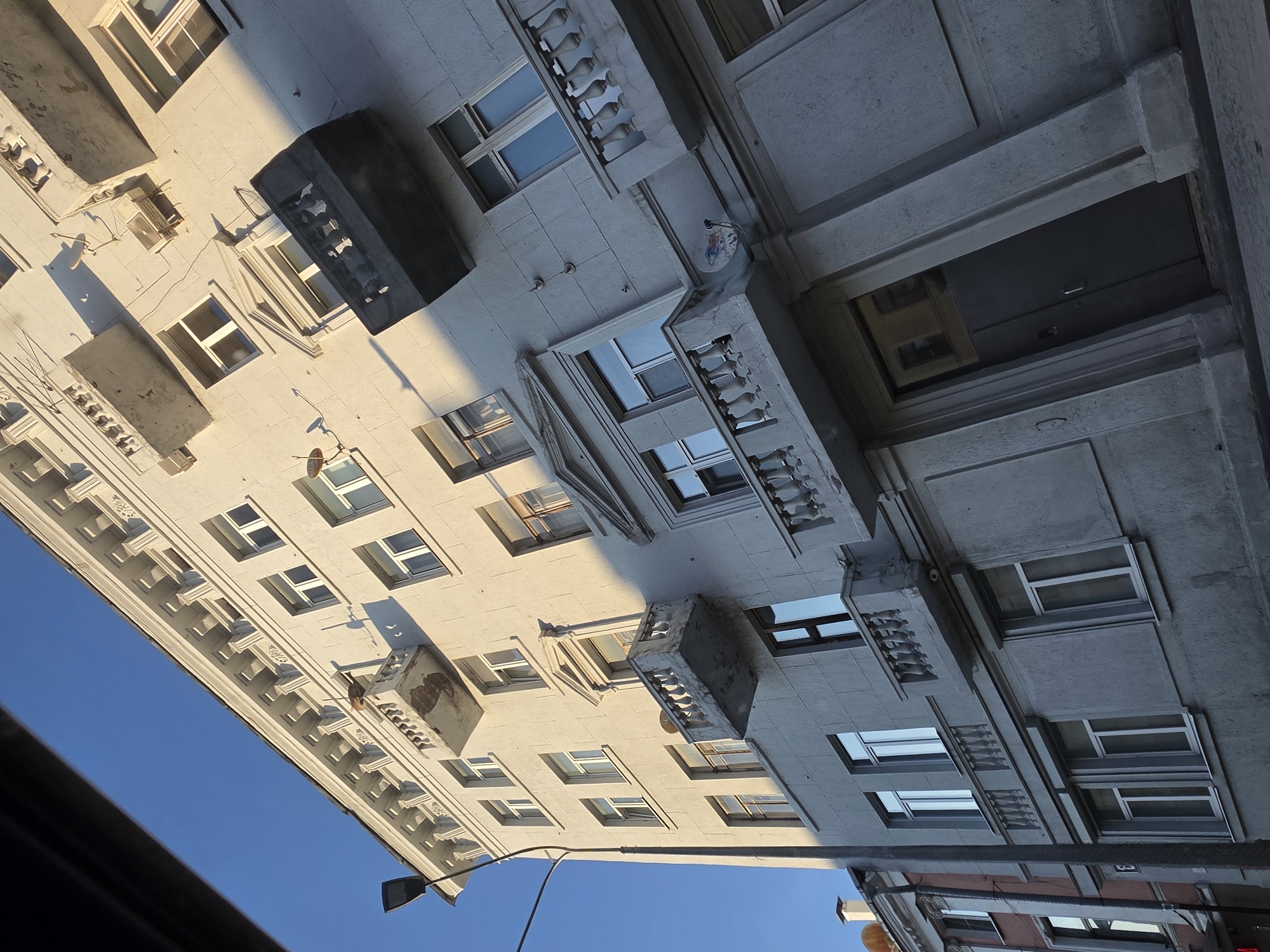 The image shows a building facade on a sunny day. The building appears to be old, with a light-colored facade and several balconies. The sun is casting long shadows across the building, indicating it is either early morning or late afternoon. There are several windows visible, some of which are reflecting the blue sky. A street lamp is visible on the left side of the image. The building has an ornate cornice at the top and some decorative elements around the windows and balconies. The perspective is from a low angle, looking up at the building. There are no people visible in the image. The building is located in the city of Nizhny Novgorod, Russia.