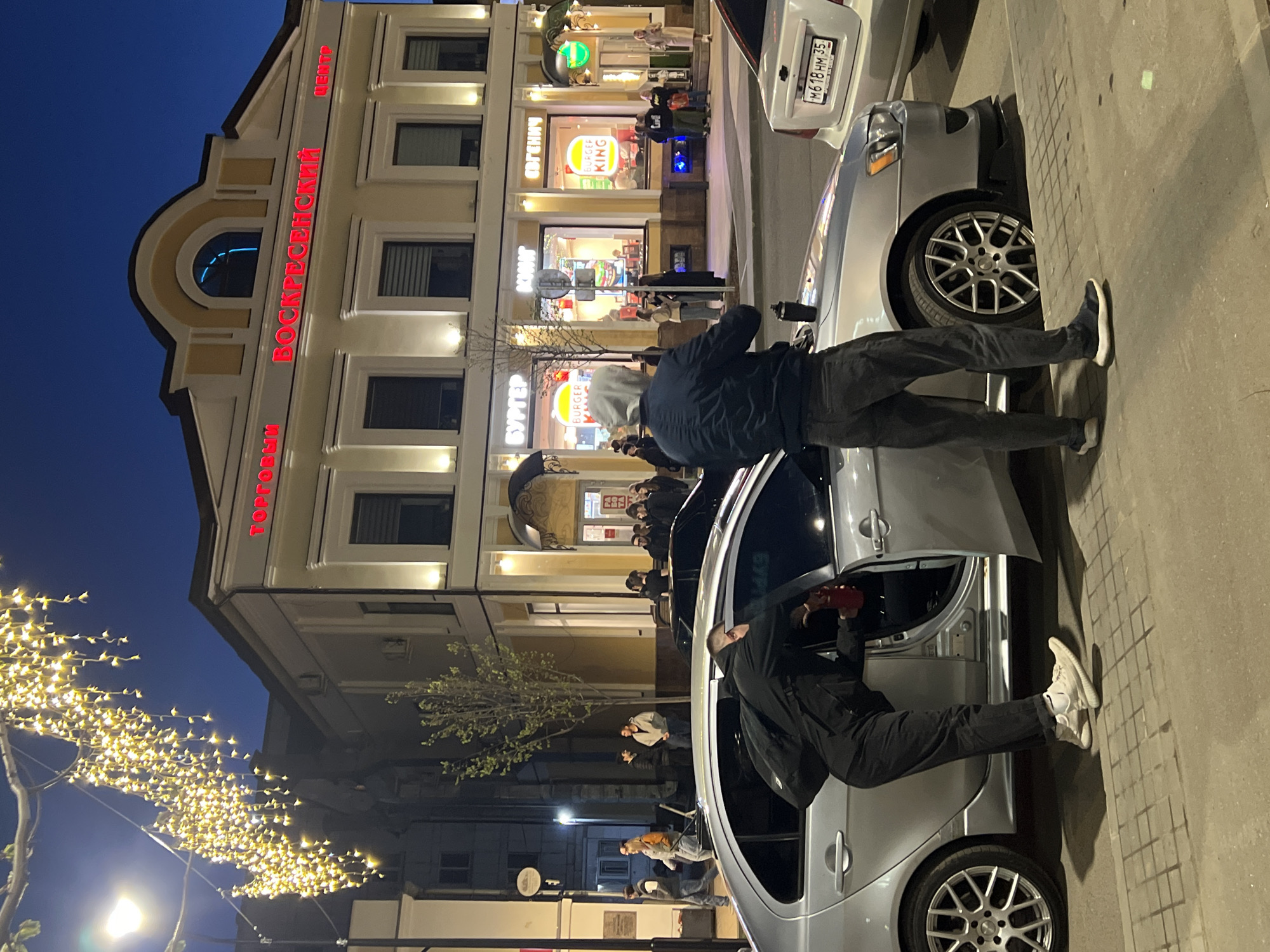 The photograph shows a street scene at night in Cherepovets, Russia. Two men are interacting with a silver car, with one man entering the driver's side and another standing outside the passenger side. The building in the background has a sign in red that reads "ВОСКРЕСЕНСКИЙ". A Burger King restaurant is visible in the background as well. Pedestrians are walking on the sidewalk. A string of lights is strung across the street. A license plate on another car reads "M618 HM 35". The lighting suggests it is dusk or early evening.
