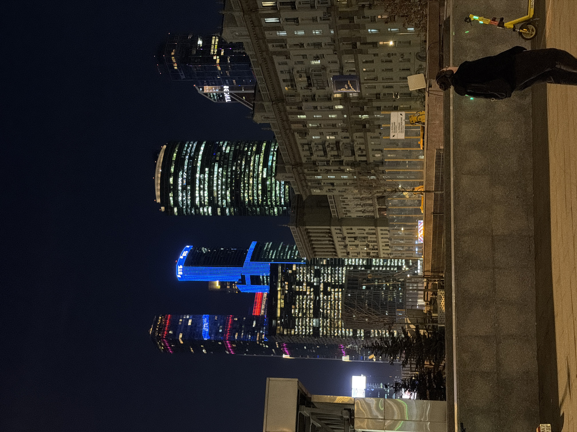 The image shows a night scene in Moscow, Russia. Several skyscrapers are visible against the dark sky. The buildings are illuminated, showcasing their architectural designs and colorful lights. A person is standing next to a wall with a yellow scooter nearby.