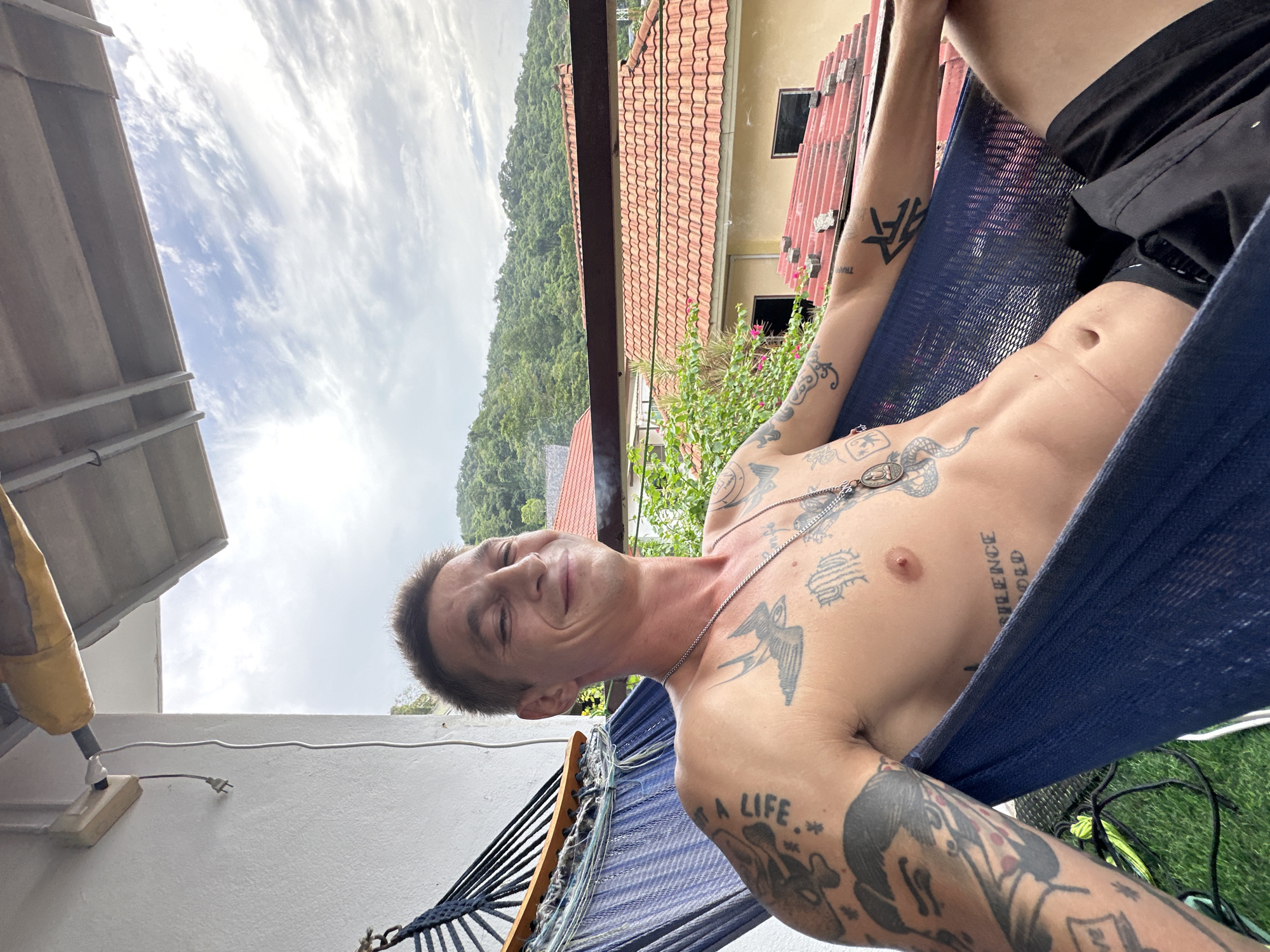 A shirtless man with tattoos is relaxing in a blue hammock on a balcony overlooking a tropical landscape. The man is smiling and looking towards the camera. The background shows lush green hills with red-tiled roofs of houses. The sky is partly cloudy. A power cord with a plug is visible on the white wall to the left of the frame. The scene appears to be in Karon, Thailand, suggested by the tropical vegetation, architecture, and the overall atmosphere.