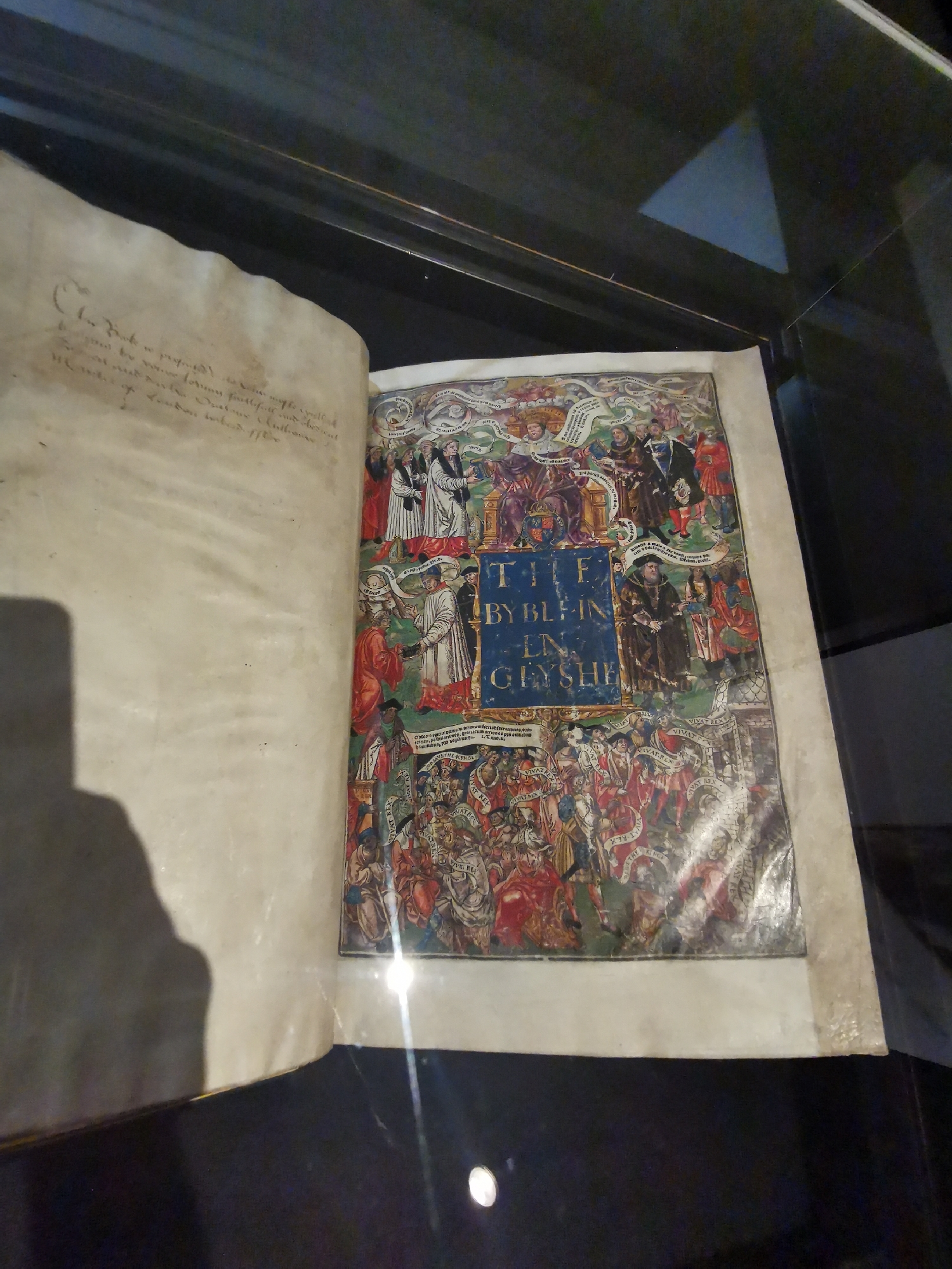 An open, large, antique book, likely a Bible, is displayed under glass in what appears to be a museum or archive setting in London, United Kingdom. The book's right page features a highly elaborate, colorful woodcut illustration, possibly a title page for an early English Bible. The central panel of this illustration prominently displays the title "THE BYBL IN EN GLYSHE" (The Bible in English).
The illustration is rich with figures and activity. At the very top, a depiction of God is visible, extending scrolls. Below this, on either side of the central title, are multiple historical figures, including kings (one with a crown and scepter), noblemen, and religious figures (bishops or monks), some holding books or receiving scrolls. These figures are arranged in a hierarchical manner, with scrolls bearing text, though largely illegible, emanating from them. Towards the bottom, large crowds of people are depicted, with many holding scrolls that clearly state "VIVAT REX" (Long Live the King), suggesting popular approval or celebration.
The left page of the book contains faded, handwritten text in an old cursive script, appearing to be an inscription or commentary. Partial legible phrases include "Chis boke is properly...", "not be found", and "faitfull and obedient".
The setting is dark, with reflections and glares on the protective glass, and a distinct shadow of a person leaning over the book is cast onto the left page. The book itself shows signs of age, with yellowish pages and some wear. The scene captures the historical significance of the book and its presentation for public viewing.