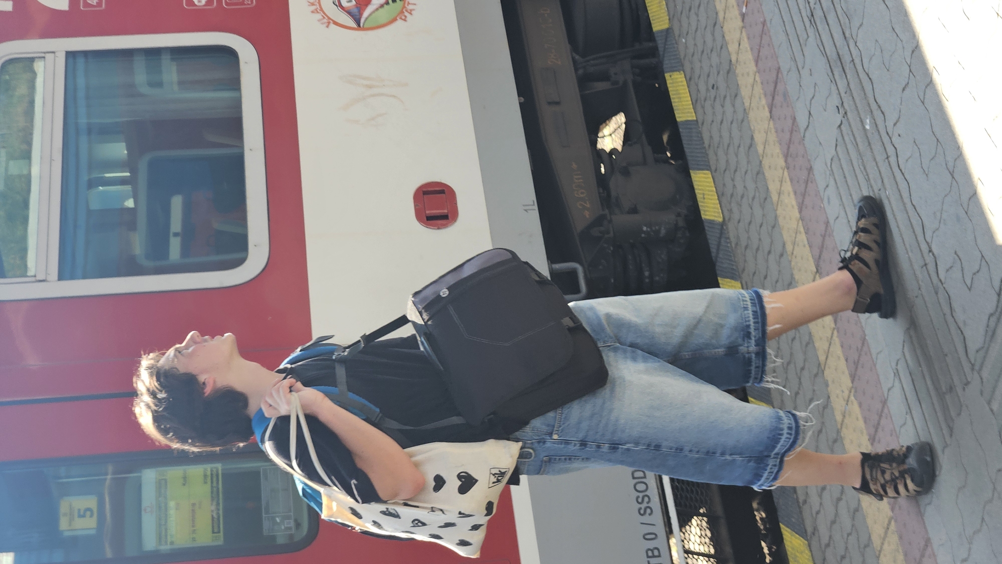 A young man with dark hair is standing on a train platform in Bratislava, Slovakia. He is wearing a black t-shirt, blue denim shorts, and brown and black sandals. He is carrying a white tote bag with black heart designs over his left shoulder and a dark messenger bag over his right shoulder. He is looking up and to his right, towards a red and white train. The train is stopped at the platform, and its windows reflect the sky. The platform is paved with gray and colored rectangular tiles, with a yellow and black striped line along the edge. The background shows the side of the train and the sky.