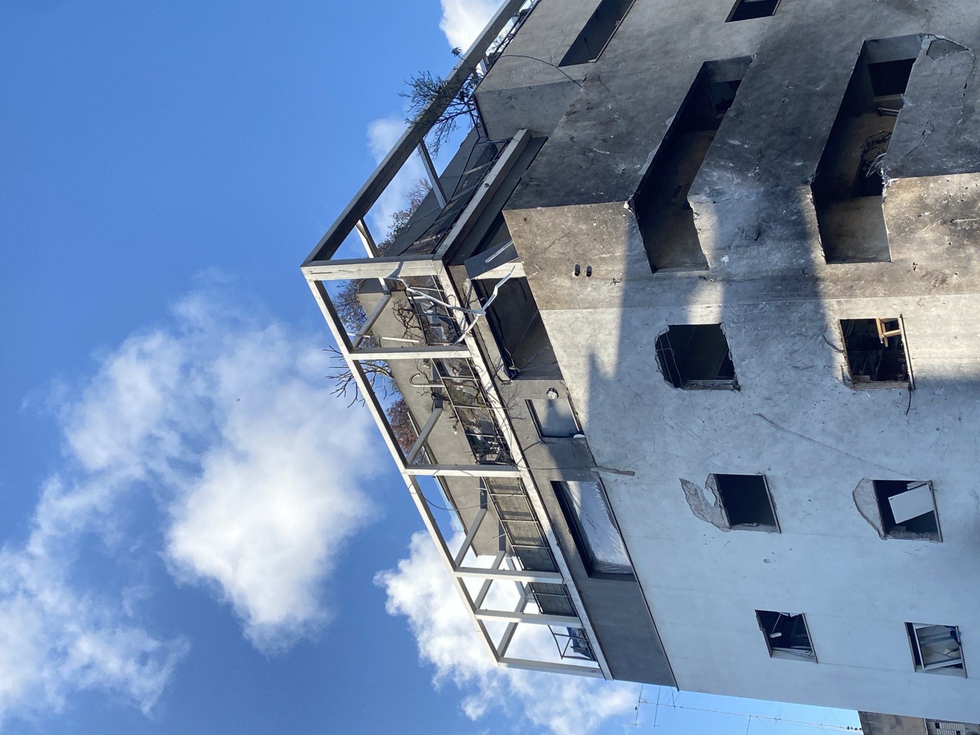 An upward-angled shot showcases a severely damaged building against a bright blue sky with scattered white clouds. The building's facade is a mix of light gray and darker, charred sections. Several window openings are visible, some with broken edges, missing glass, and interior debris. A skeletal framework of metal beams projects outwards from the building, supporting what appears to be remnants of greenery, possibly overgrown plants or small trees. The building's overall condition suggests significant structural damage, possibly from a fire or explosion. The location appears to be Tel-Aviv, Israel, based on the architectural style and urban setting. The image portrays a scene of destruction and urban decay.