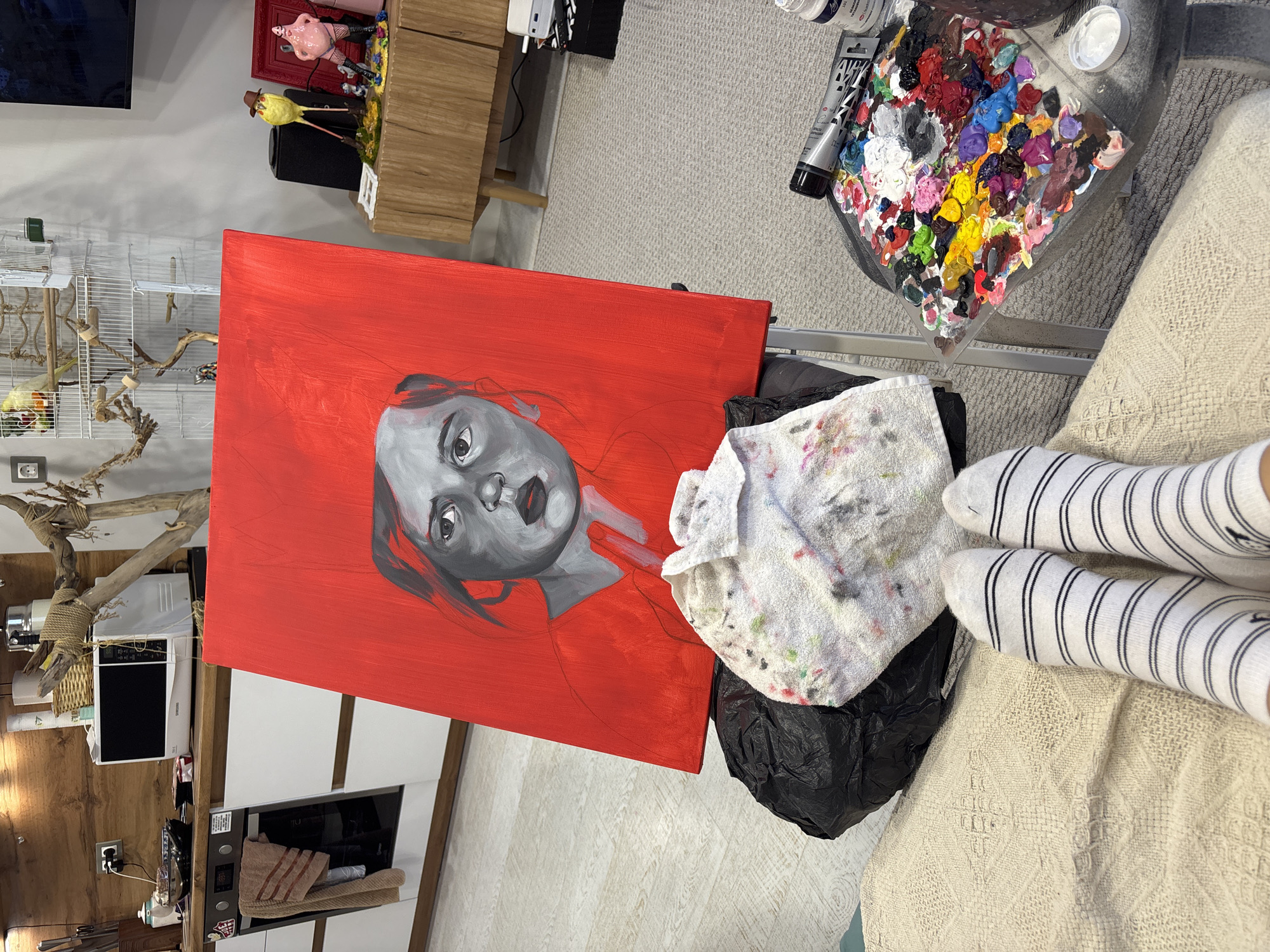 This image captures a relaxed indoor setting where an individual is engaged in painting. A pair of feet, clad in white socks with thin black horizontal stripes, rest comfortably on a textured, light-colored blanket, suggesting the person is seated on a sofa or armchair.
In the foreground, a large canvas displays an in-progress portrait. The painting features a grayscale face, possibly of a woman, rendered with striking detail on the eyes and expressive lips, set against a vibrant, solid red background. Beside the canvas, a clear plastic palette holds a diverse collection of dried and wet acrylic paints in numerous colors, along with paint tubes, one visibly labeled "ART ACRYLIC." A paint-stained white towel is also on hand, indicating active work.
The background reveals parts of a home environment. To the left, a white birdcage contains a light-colored parakeet or budgie perched on a branch. Nearby, a wooden cabinet features a black speaker topped with a whimsical yellow bird figurine wearing a cowboy hat, and a red framed picture with a pink figurine. Further back, kitchen elements such as a "Samsung" microwave and an oven integrated into white cabinets are visible, alongside wooden wall paneling and what appears to be a light-colored carpet and wooden flooring. The scene suggests a peaceful, creative moment taking place indoors.