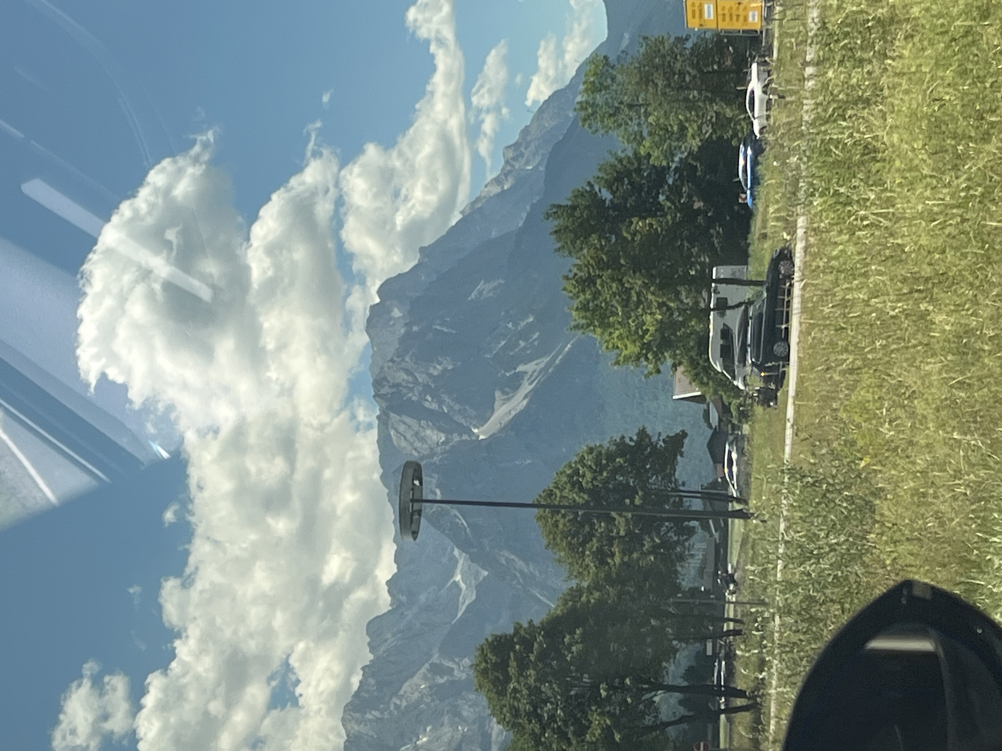 The image shows a scenic view of the Alps, likely the Zugspitze massif, from the vicinity of Garmisch-Partenkirchen, Germany. In the foreground, there is tall, dry grass and a portion of a car's side mirror. Behind the grass, a road can be seen with several cars, including a black SUV, a blue sedan, and a white sedan. Further back, there is a white camper van and a tall, industrial-looking pole with a circular top. Behind the road and vehicles are several large, mature trees. The majestic, snow-capped mountains of the Alps dominate the background, with a clear blue sky dotted with fluffy white clouds above. The overall impression is one of a bright, sunny day in a mountainous region, with signs of human presence in the form of vehicles and infrastructure.