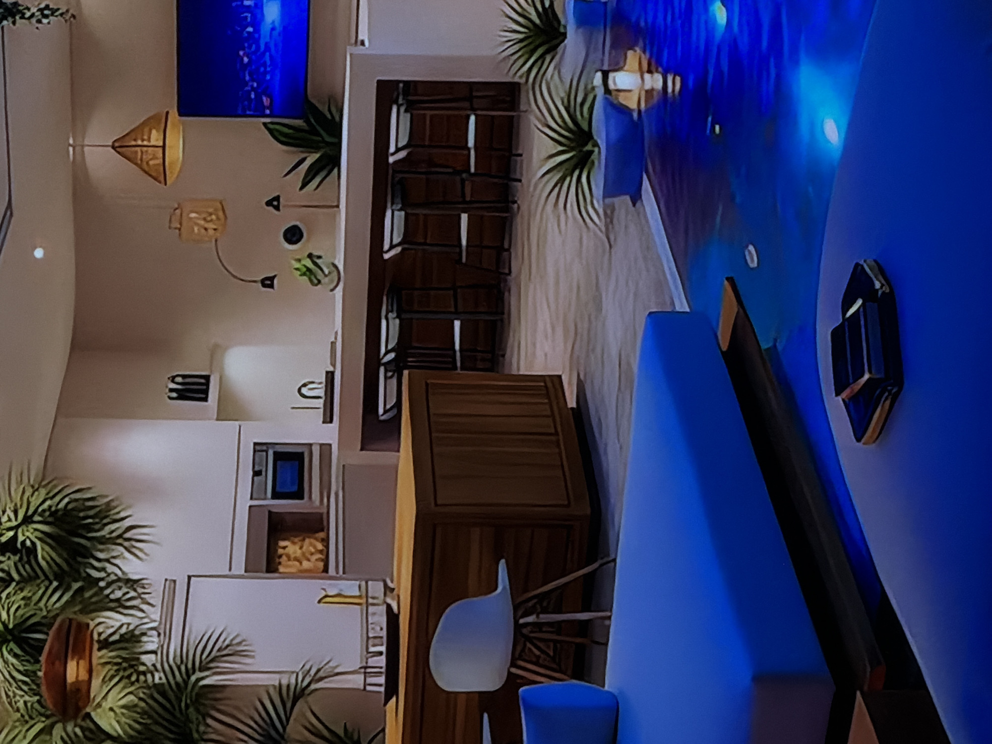 The image displays a modern, upscale interior space, appearing to be an open-plan kitchen and living or dining area. The overall aesthetic is characterized by a vibrant blue and warm wood color palette, accented with lush greenery.
In the midground, a contemporary kitchen features light-colored, possibly white, cabinetry with a built-in microwave or oven and a stainless steel faucet over a sink. Adjacent to the kitchen is a long wooden bar or island, lined with several dark-legged bar stools. Overhead, two distinctive pendant lights, one triangular and one rectangular, both crafted from a light, woven material, hang from the ceiling. A unique floor or wall lamp with multiple spherical lights on dark metal arms is also visible.
Towards the right side of the frame, a large, round table with a striking blue surface occupies the foreground, accompanied by a matching blue sofa or bench seating. A modern light-colored chair with wooden legs is also present near a wooden cabinet or sideboard. Behind the bar area, a large wall-mounted screen or illuminated art piece displays vivid blue, abstract patterns, casting a glow and reinforcing the room's dominant color.
Several large, green potted plants, resembling palms or ferns, are strategically placed, including a prominent one in the bottom left foreground and others near the bar and seating areas, adding a natural and calming element. The space is brightly illuminated, suggesting either daytime or a well-lit evening. There are no people visible, indicating an unoccupied setting. No specific external location cues or text can be discerned within the image itself.