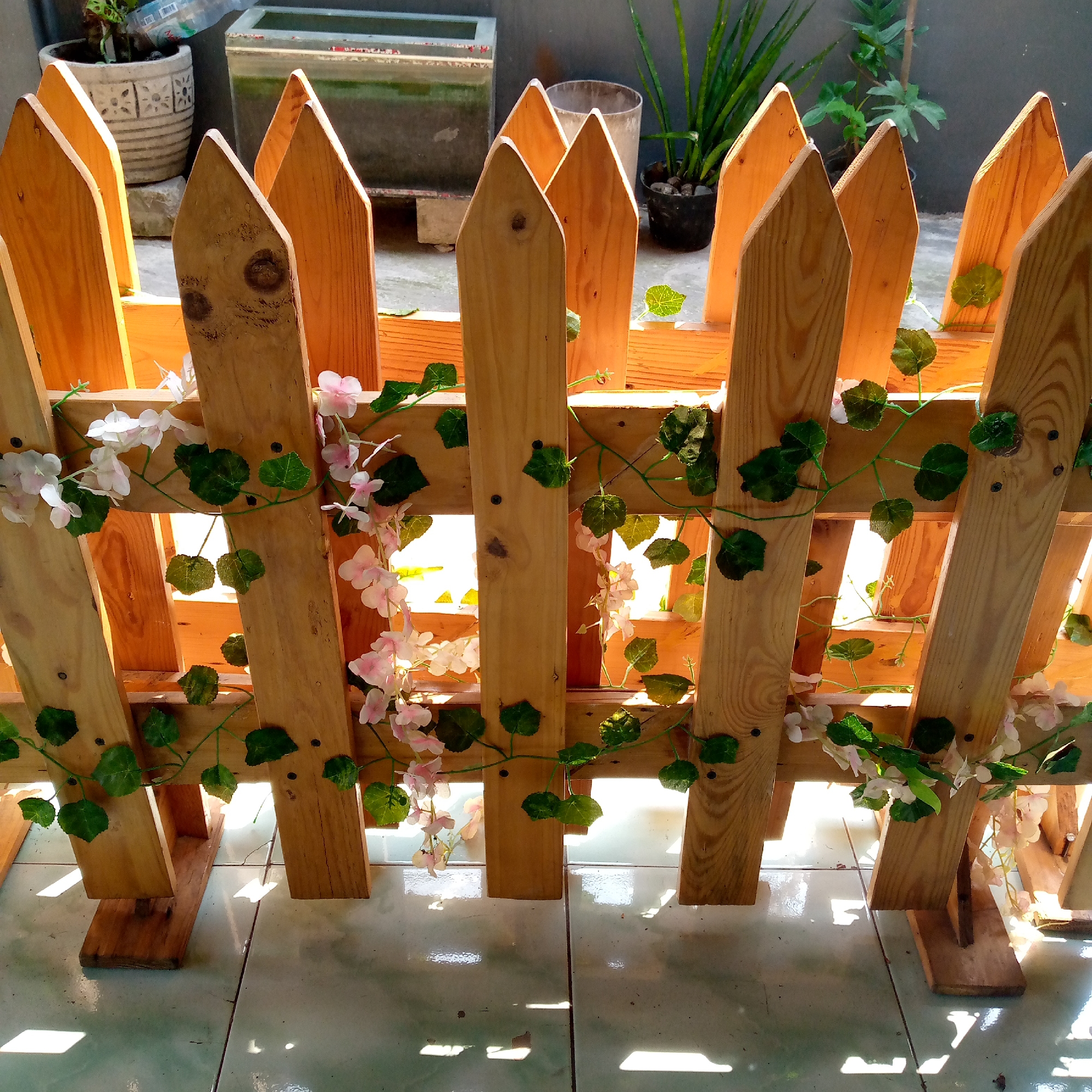 The image shows a decorative wooden fence with artificial flowers and vines. The fence is made of pointed wooden planks, stained a light brown. Green artificial leaves and small pink and white artificial flowers are intertwined with the vines, which are wrapped around the fence. The fence is situated outdoors on a tiled surface, and sunlight is casting shadows through the gaps in the fence. In the background, out of focus, are some potted plants and a grey wall. The scene appears to be a residential outdoor area, possibly a yard or garden. The specific location in the city of Sumberwono, Indonesia, is not discernible from the image itself.