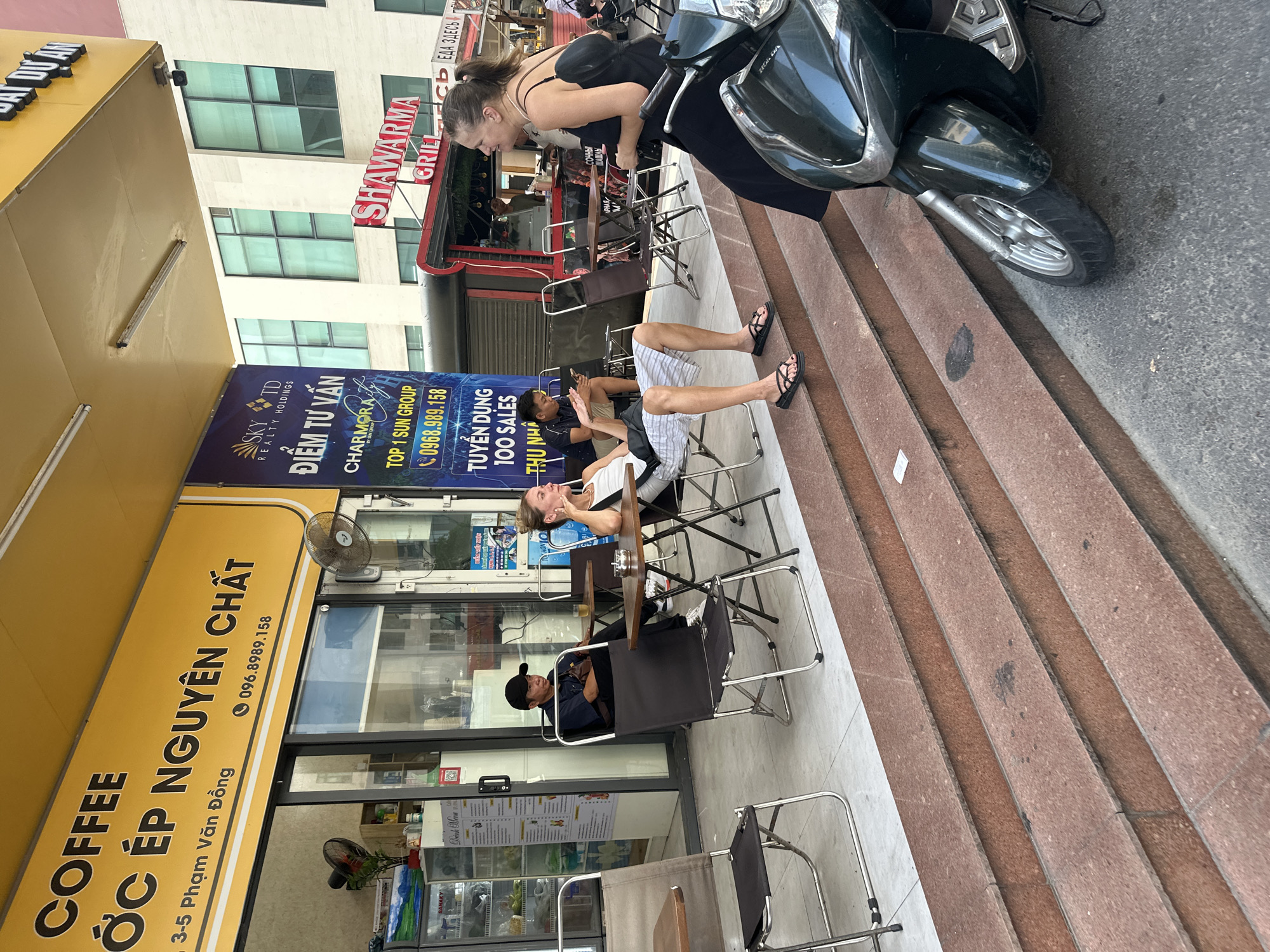 Here's a concise description of the image:
The scene is set in Bac Nha Trang Ward, Vietnam, outside a "COFFEE ỚC ÉP NGUYÊN CHẤT" shop at 3-5 Phạm Văn Đồng. A woman in a black dress is leaning over a dark green scooter parked on a set of stairs. Below, two people are sitting at tables outside the coffee shop. One person is lounging with their legs extended, while the other, wearing a cap, sits upright. Behind them, a banner advertises real estate and job opportunities. In the background, a restaurant named "SHAWARMA GRILL" can be seen. The environment is urban with storefronts and pedestrian activity.