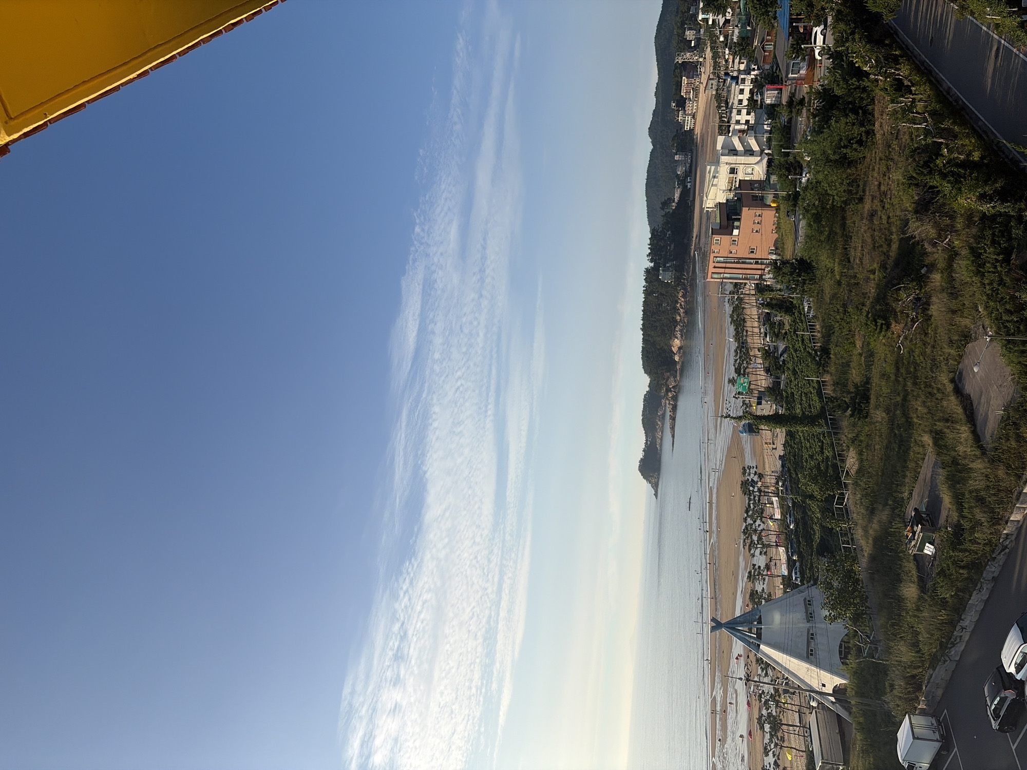 The image shows a coastal scene in Mohang-ri, South Korea, likely from an elevated vantage point. In the foreground, there is an area with overgrown grass and vegetation, a road, and parked vehicles, including a white truck and several cars. A distinctive, cone-shaped building, resembling a teepee, is situated on the left side of the frame. Beyond this, a sandy beach is visible with scattered people and umbrellas. The ocean stretches out to the horizon, meeting a pale blue sky with wispy clouds. In the distance, hills covered in trees and a small cluster of buildings line the coast. The overall atmosphere is bright and appears to be during daylight hours.
