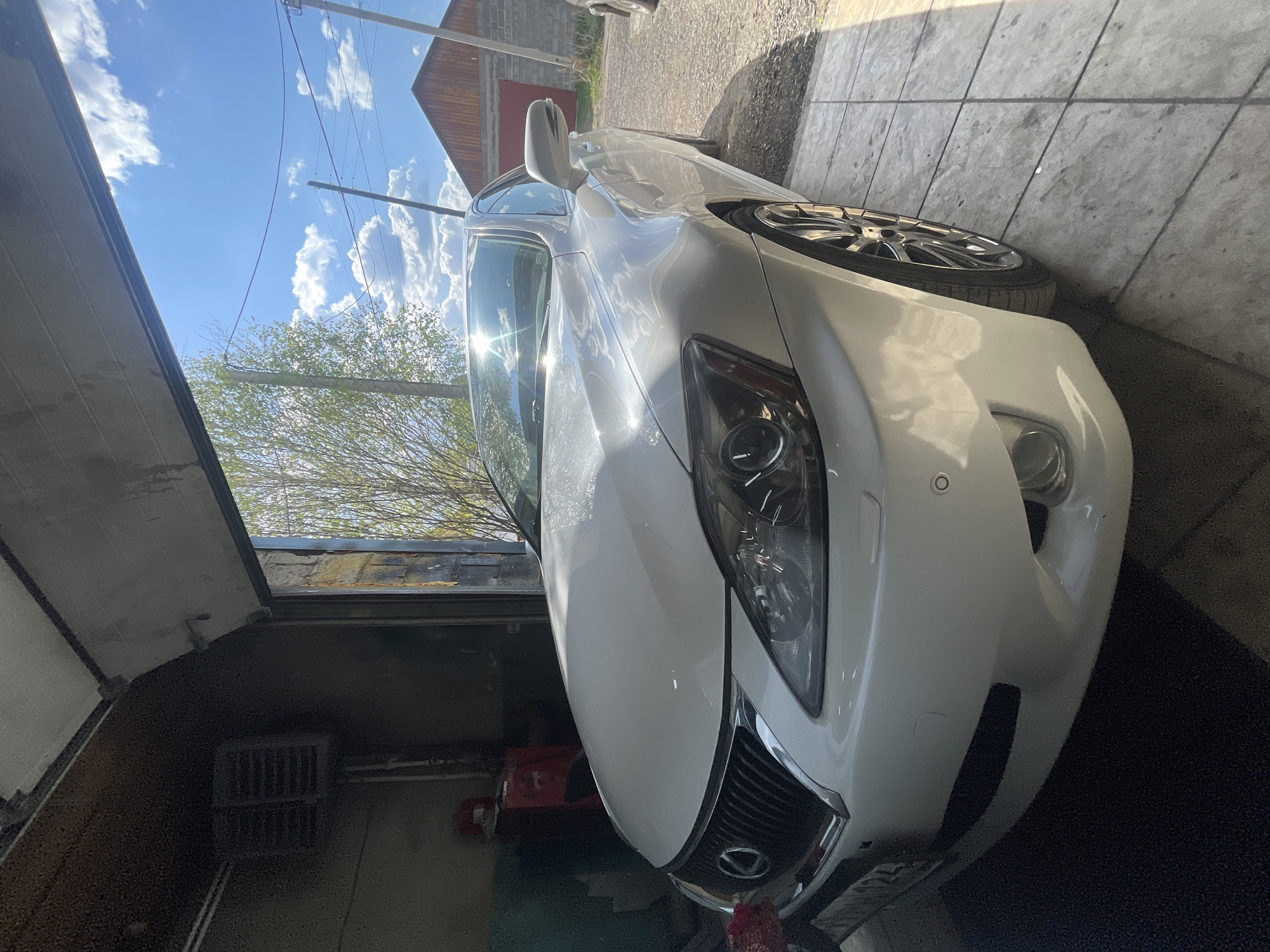 The image shows a white car parked in a garage in Минино, Russia. The front of the car faces the camera, and the garage door is partially open, revealing a view of the sky and trees outside. The car is positioned close to a wall with stone paving. There are no people visible in the frame.