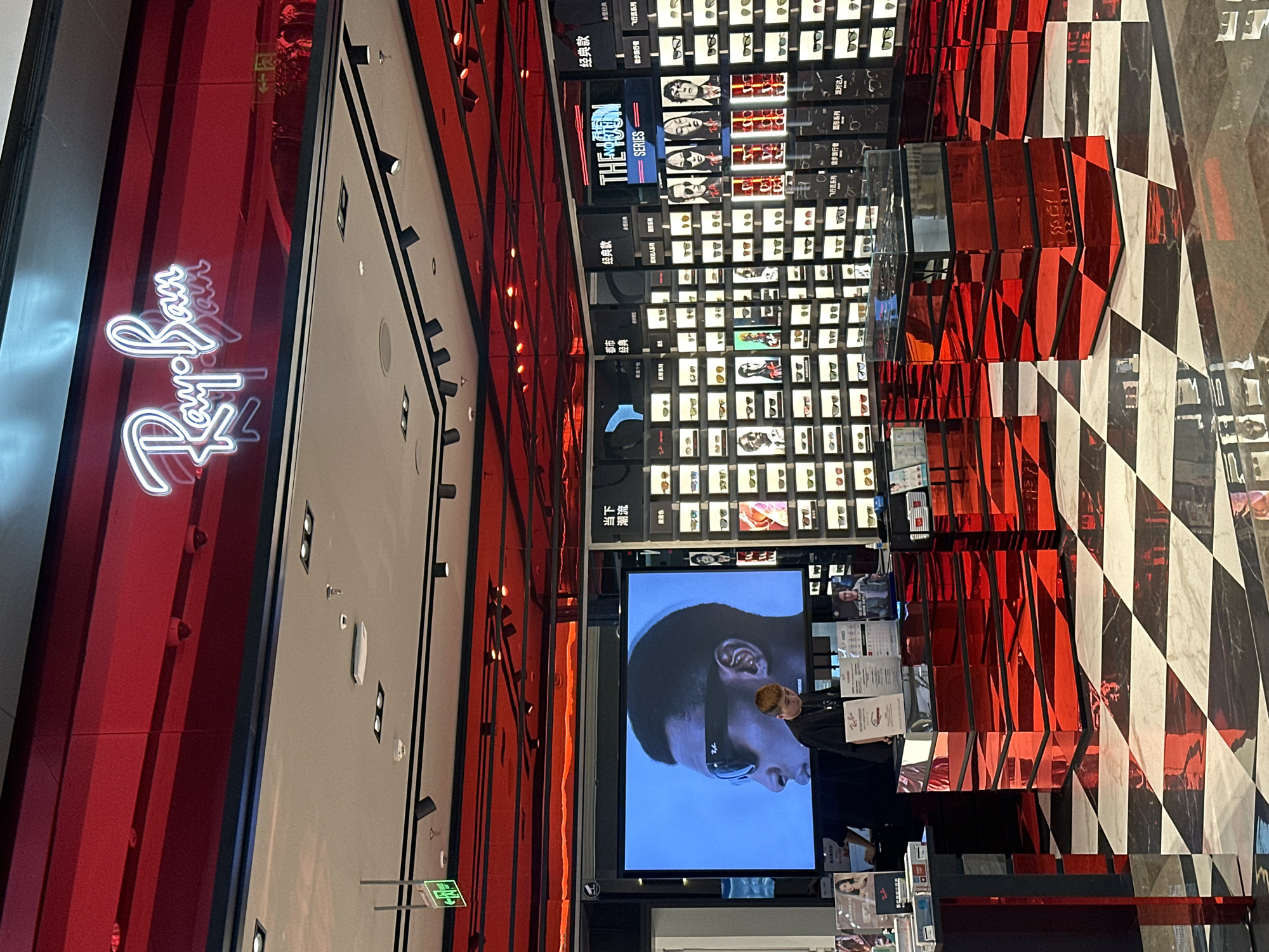 Here is a description of the image:
The image depicts the interior of a Ray-Ban store, likely in Liangjiang, China, due to the presence of Chinese characters on signage. The store's design is striking with a red and black color scheme. The prominent Ray-Ban logo is visible in neon lighting. Display cases are filled with various Ray-Ban sunglasses. A large screen displays an image of a person wearing sunglasses. The floor is a checkerboard pattern of black and white tiles. A sales associate is standing behind a counter. The general atmosphere suggests a modern retail environment.