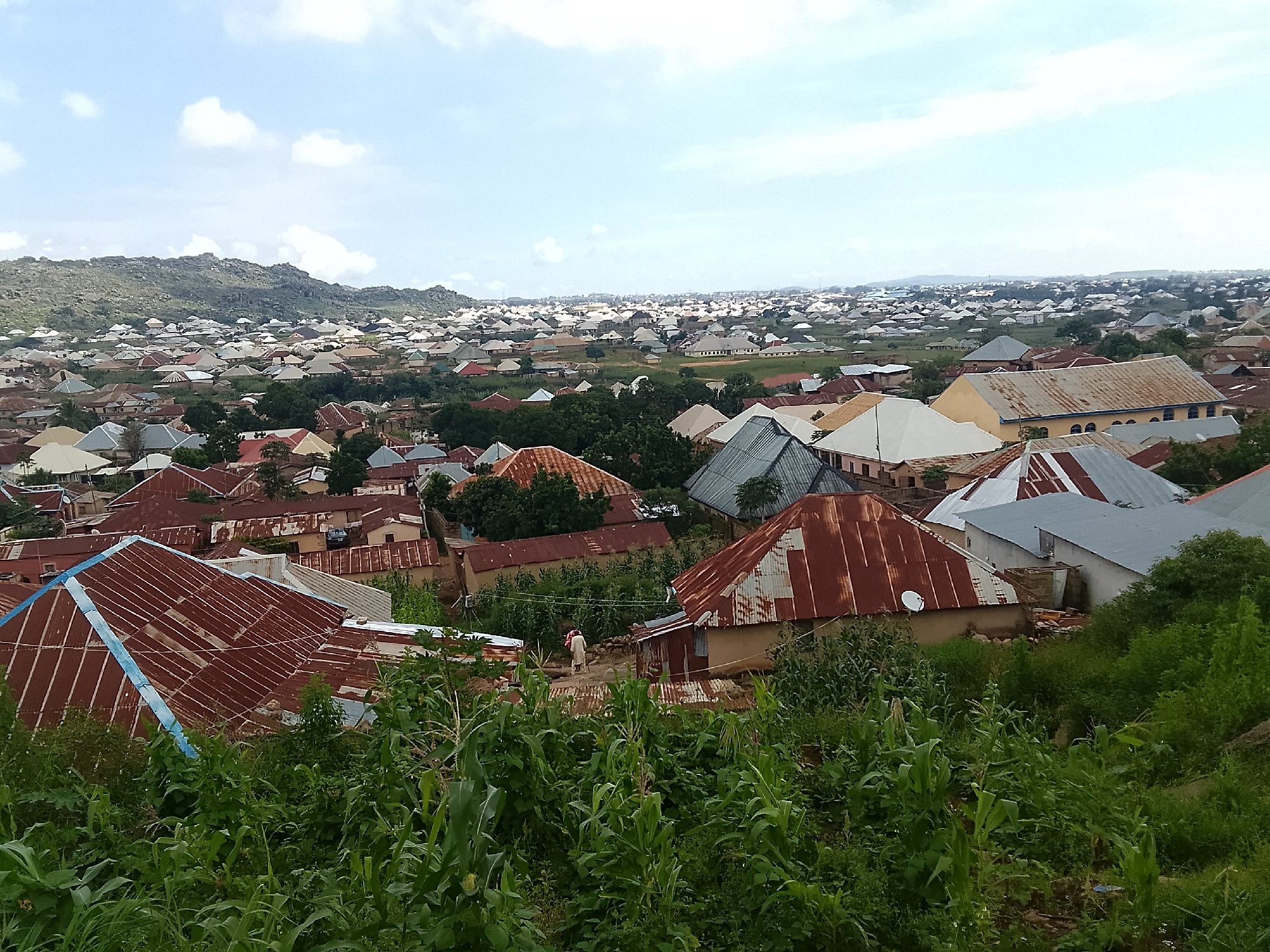 This is a wide-angle, eye-level shot of a densely populated residential area in Rukuba Road, Nigeria. The foreground is dominated by lush green vegetation, primarily tall corn stalks. In the mid-ground and background, a sprawling collection of houses with corrugated metal roofs in various states of repair, ranging from rusty red to dull gray, are visible. Some houses have white or light-colored walls, while others have natural earth tones. The rooftops vary in shape and pitch, with some appearing flat and others steeply sloped. Scattered trees are interspersed among the buildings. In the far distance, a range of hills or mountains forms the horizon, under a pale blue sky with scattered white clouds. A few individuals are visible on a dirt path in the mid-ground, one appears to be walking with another person. The overall impression is of a vibrant, somewhat rustic urban landscape.