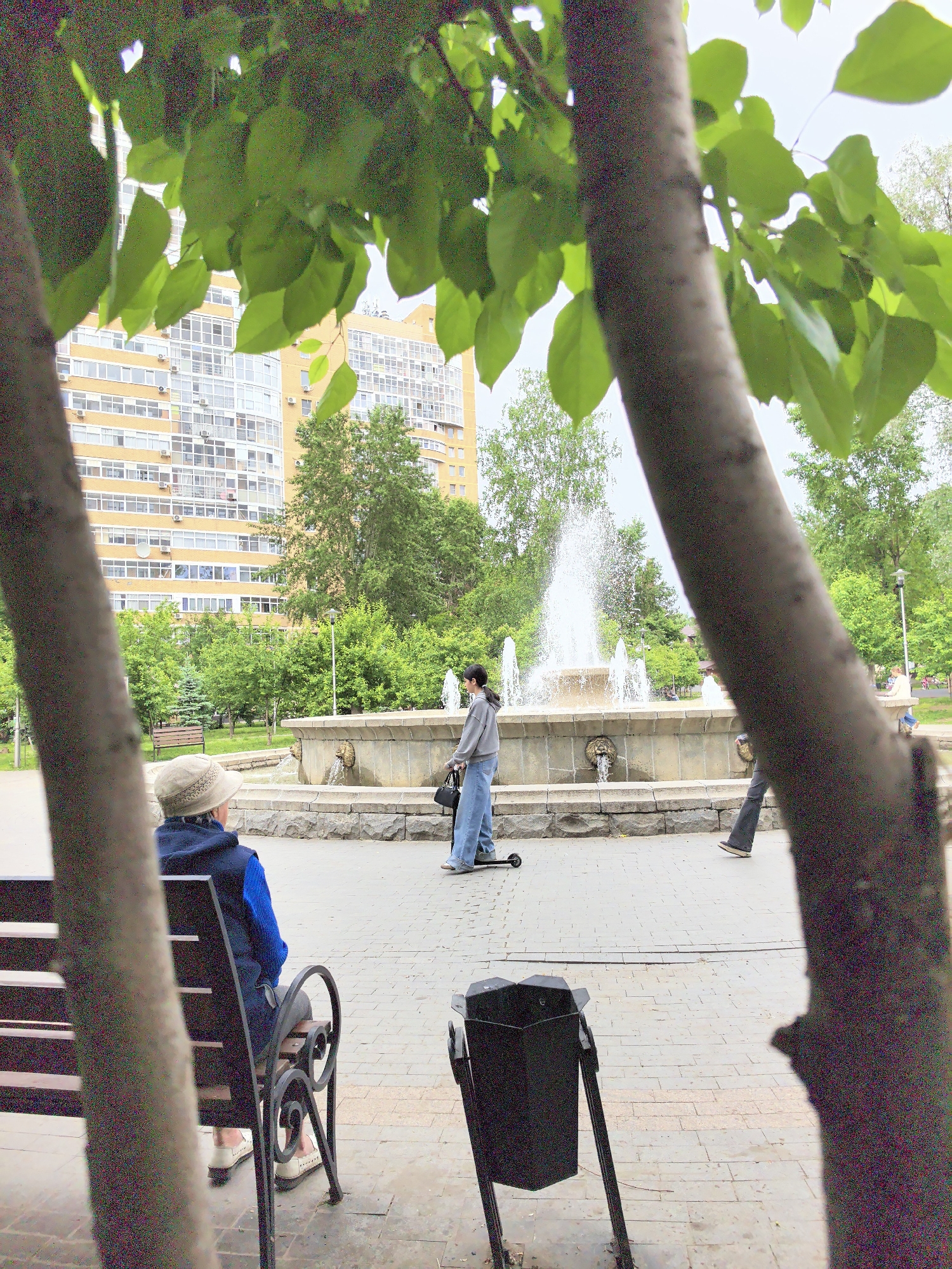 The image captures a public park or square scene, framed through the leaves and trunks of two trees in the foreground. In the background, a large, light-yellow, multi-story apartment building with numerous windows is visible.
In the midground, a stone fountain with water actively spraying upwards and spouting from decorative lion heads is the central feature. To the left of the fountain, a person wearing a light-colored hat, a blue sweater, and a dark vest is seated on a black park bench, facing away from the camera. To the right of the fountain, another person, dressed in a grey hoodie and blue jeans, stands next to a black kick scooter, also facing away. A third person's legs are partially visible walking past the fountain on the right side of the frame.
The ground is paved with light-colored bricks. Between the fountain and the apartment building, there are various green trees and bushes, suggesting a lush park environment. A black metal trash can is positioned in the lower right foreground. The lighting indicates it is daytime, possibly overcast, given the soft, diffused light. There is no visible text in the image. The general atmosphere is calm, with people enjoying a public space. The location is unknown.