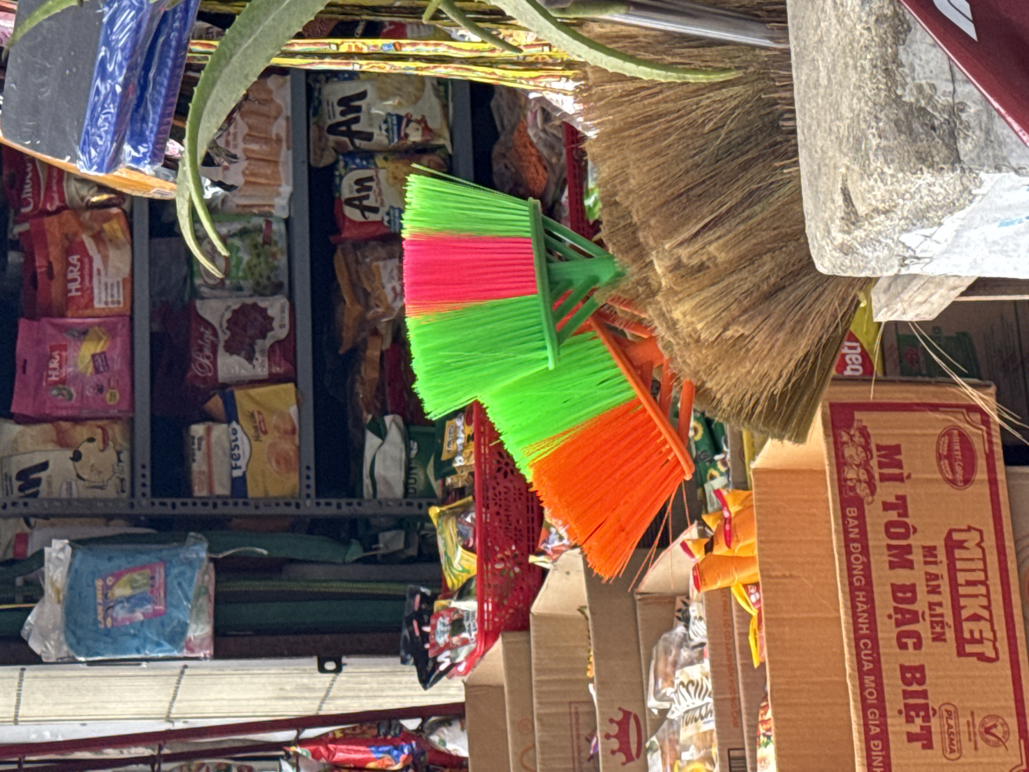 The image provides a detailed, vertical view of a crowded retail shelf or market stall in Lâm Viên - Đà Lạt, Vietnam, showcasing a variety of everyday consumer goods. No people are visible in the frame.
In the foreground, several cleaning items are prominent: two brightly colored hand brushes, one with vibrant pink and neon green bristles, and another with orange bristles. A larger, natural fiber broom or brush is also partially visible on the right.
The shelves are packed with numerous packaged food products. Among the discernible brands and items are "HURA" on pink and orange packages, "An An" on white snack bags, as well as "Festo," "Belgi," and "Chow" on other assorted packets. A large cardboard box on the right clearly displays the brand "MILIKET" and identifies its contents as "MÌ ĂN LIỀN" (Instant Noodle) and "MÌ TÔM ĐẶC BIỆT" (Special Shrimp Noodle). Additional text on this box includes "BẠN ĐỒNG HÀNH CỦA MỌI GIA ĐÌNH" (Companion for every family), "PLASMA," and "MILIKET-Copral." Other partial texts like "bati," "DUN," "S," "LANH," and "CUNG" are also seen on various packaging.
A green leafy plant or vine extends into the upper left portion of the image, partially obscuring some items. A blue packaged item, possibly a cloth or blanket, is also visible on a lower shelf. The lighting suggests an indoor or shaded environment, with no specific time of day or weather conditions evident. The arrangement conveys the bustling and practical atmosphere of a local Vietnamese market or convenience store.