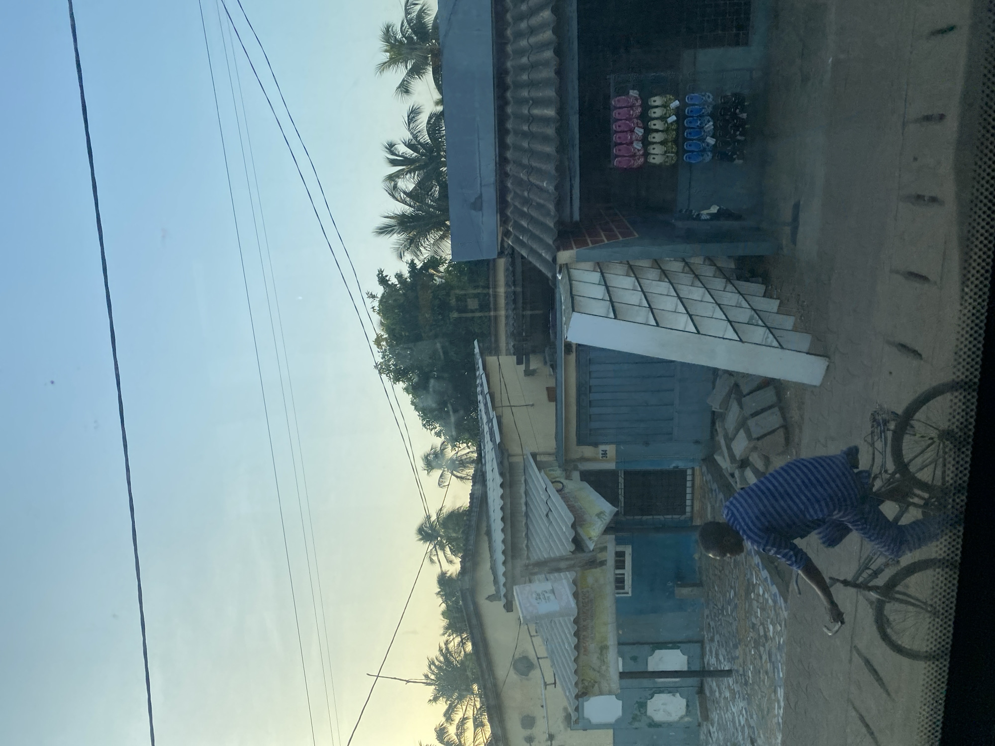 A street scene in Aného, Togo, features a man wearing a blue striped shirt riding a bicycle. Buildings are visible along the street, including a shop displaying footwear for sale. The weather appears to be clear and sunny, and palm trees are seen in the background. Overhead power lines are present. The image seems to be taken from inside a vehicle, looking out onto the street.