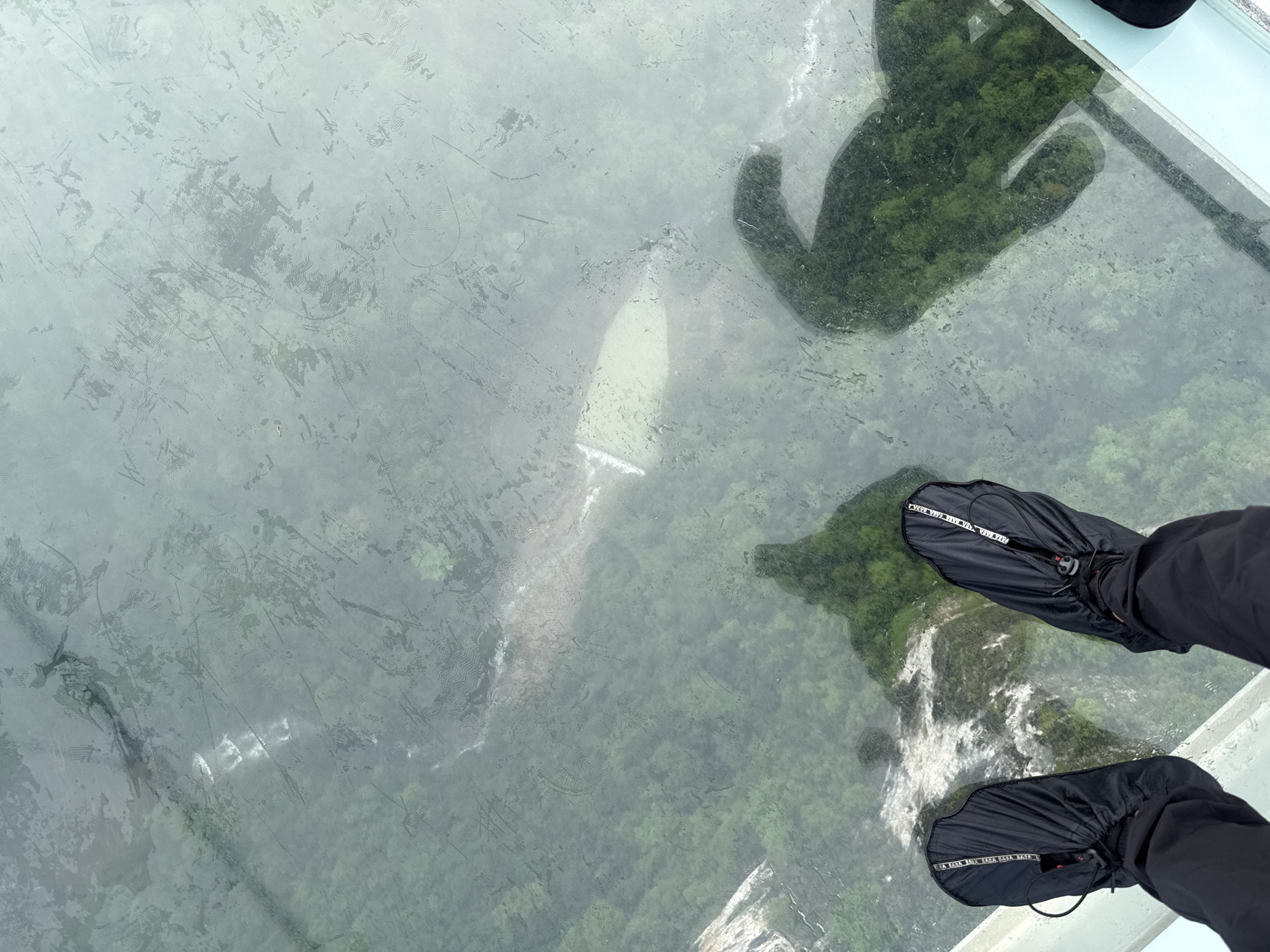 From a high-angle perspective, the image depicts a view through a glass-bottomed walkway, likely the Zhangjiajie Glass Bridge in Zhangjiajie, China. Below the glass, dense green foliage and rocky terrain are visible, creating a sense of height and depth. The glass surface is somewhat obscured by what appears to be light scratches, water spots, or condensation, which slightly blurs the view below. Two feet encased in black protective shoe coverings with visible straps are in the lower right of the frame, indicating the viewer is standing on the glass. Overlaid on the glass are the shadows of people standing on the bridge.