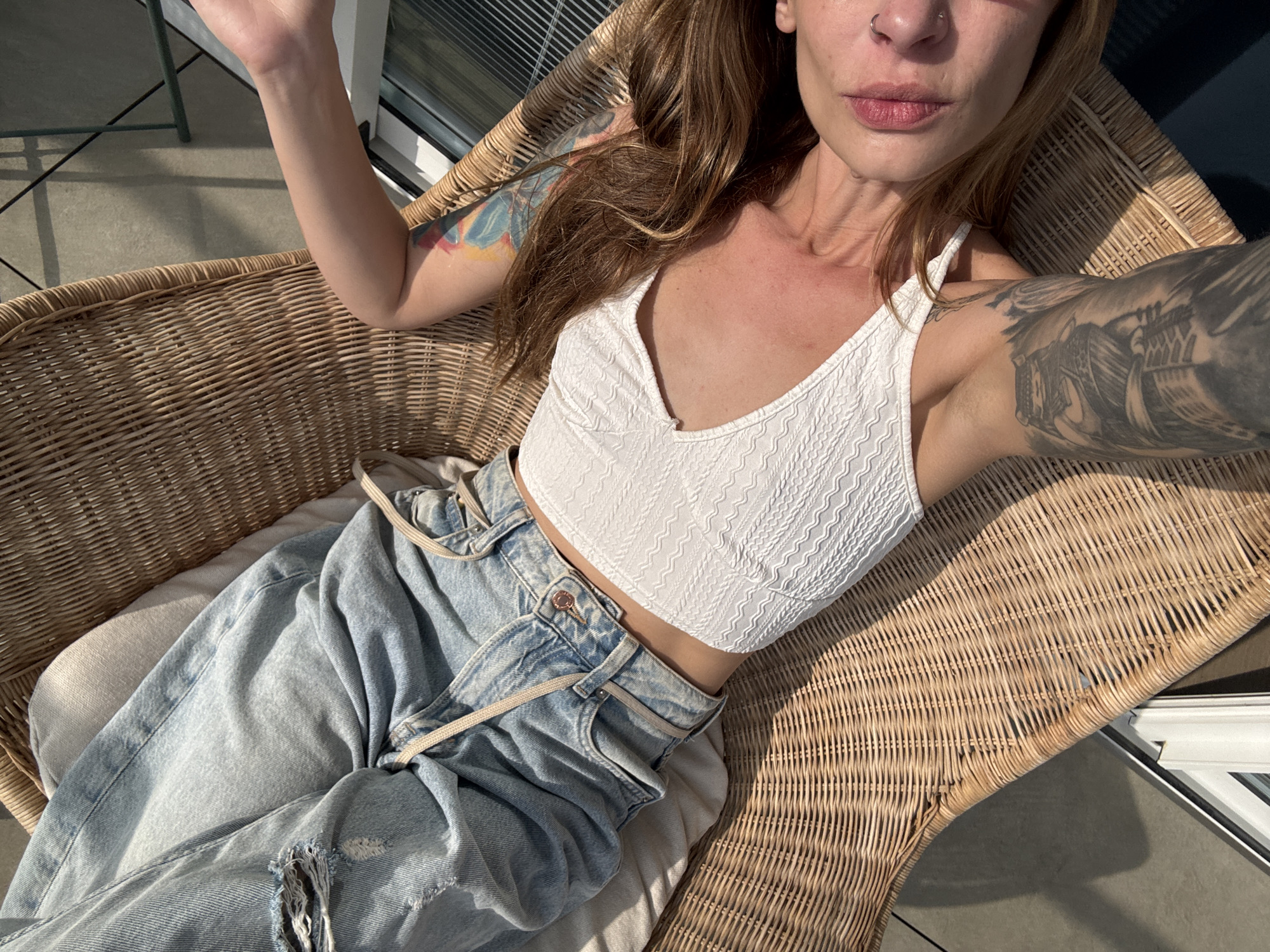 A woman with tattoos lies in a wicker chair outdoors. She wears a white crop top and light wash jeans with a drawstring and a hole at the knee. Her arms are raised, one slightly bent. The chair is positioned near a window or balcony door. Based on the style of architecture and the presence of blinds and a wicker chair, the location could potentially be a private balcony or patio in Tel-Aviv, Israel.