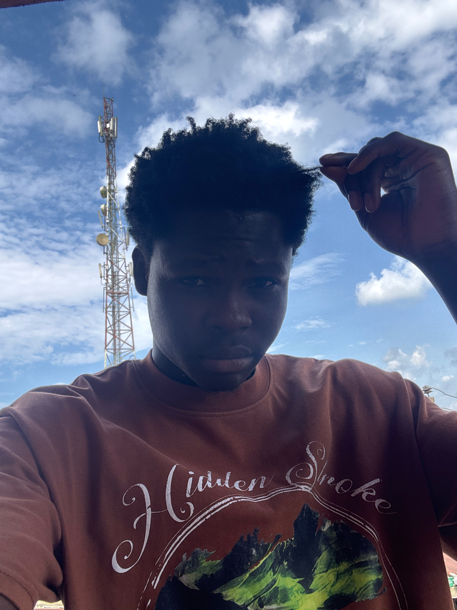 A young Black male with dark, curly hair is pictured outdoors. He is wearing a reddish-brown t-shirt with white text that reads "Hidden Smoke" and a graphic of a mountainous landscape. His right hand is raised, and he is touching the ends of his hair. The background features a bright blue sky with scattered white clouds. A tall telecommunications tower is visible behind him, to his left. The image appears to be taken from a slightly low angle, looking upwards.
