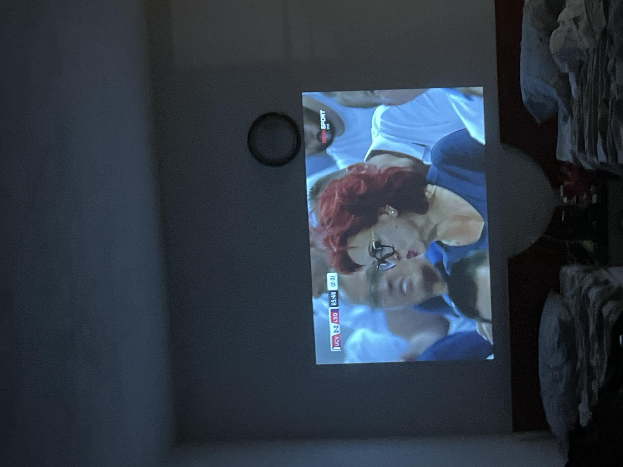 An indoor scene depicts a projection of a sports broadcast onto a wall, possibly in a bedroom given the presence of beds in the foreground. The projected image shows a woman with bright red hair and glasses in the foreground, and behind her, several people with blurred faces are visible. A scoreboard at the bottom left of the projected image indicates "UCV 2-2 LSO 85:48 (2-3)". The top right corner of the projection displays a logo that reads "SPORT LIVE". The room is dimly lit, with the projector casting light onto the wall. The overall impression is of a person or people watching a game from a relaxed setting. The specific location in Obzor, Bulgaria, cannot be determined from the provided image.