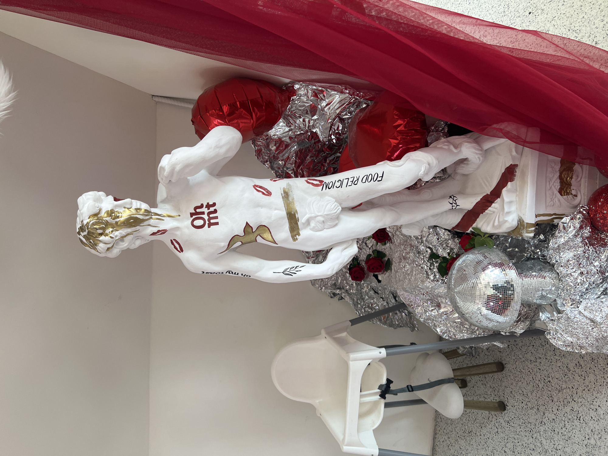A customized replica of Michelangelo's David statue is displayed in an indoor setting. The white statue features gold paint dripping from its head and face, red lipstick kiss marks on its chest and upper thighs, and various text and symbols. "Oh mt" is written in bold red letters on the chest, a golden bird figure is on the abdomen, and a horizontal gold stripe is below it. "On my toast" is written vertically on the statue's left upper arm, and a black stylized leaf or branch is on its lower left arm. "FOOD RELIGION" is written vertically on the right thigh. A red band, appearing to be paint, is around the lower right leg.
The statue is surrounded by decorative elements. To its right and behind it, a pile of crumpled silver foil material is intermixed with several red Mylar balloons. Three red roses are visible on the silver foil near the statue's base on the left. In the lower left foreground, a silver disco ball shaped like a mushroom rests on the silver foil. A large piece of sheer red fabric hangs vertically along the right side of the frame, partially obscuring the background.
The background consists of plain white walls. To the far left, a white high chair with gray legs and a black strap is visible. The floor is light-colored with a speckled, possibly terrazzo, pattern. No people are present in the scene. The location in the city of Moscow, Russia cannot be determined from the visual evidence provided.