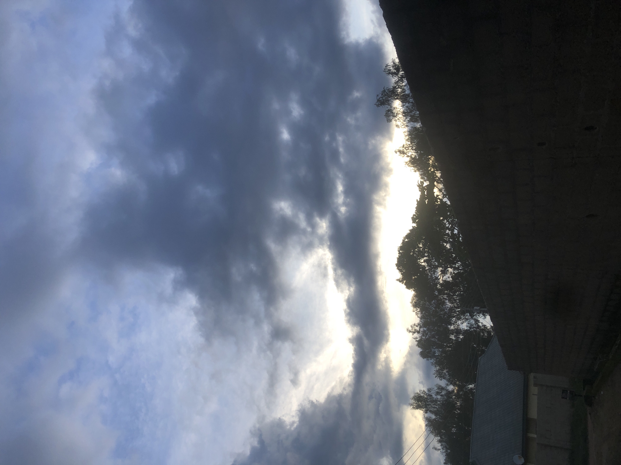 The image depicts a scene in Barakin Lerawi, Nigeria, under a dramatic sky filled with dark, heavy clouds partially illuminated by a bright light breaking through near the horizon. In the foreground, a rough, dark brick wall dominates the right side of the frame, casting a deep shadow. To the left, the roof of a building with a metallic, grey finish is visible, with a satellite dish mounted on its side. Beyond the buildings, a line of dense trees with thin branches forms a dark silhouette against the bright sky. The overall impression is one of an approaching storm or the aftermath of one, with the natural light struggling to pierce the thick cloud cover. The environment appears to be a residential or semi-rural area. There are no people visible in the image.
